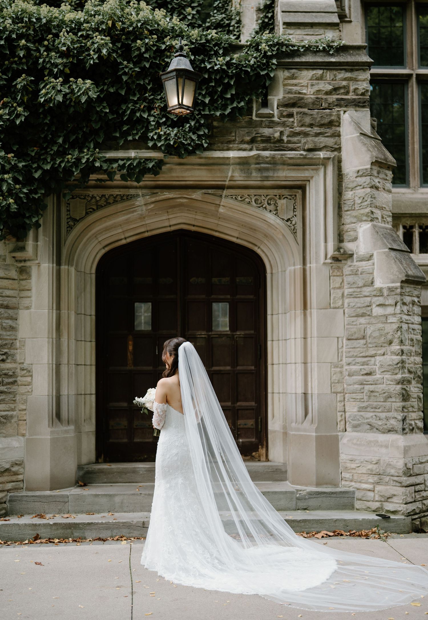 A long flowing cathedral veil trails behind on stone steps in front of a Gothic-style church entrance.
