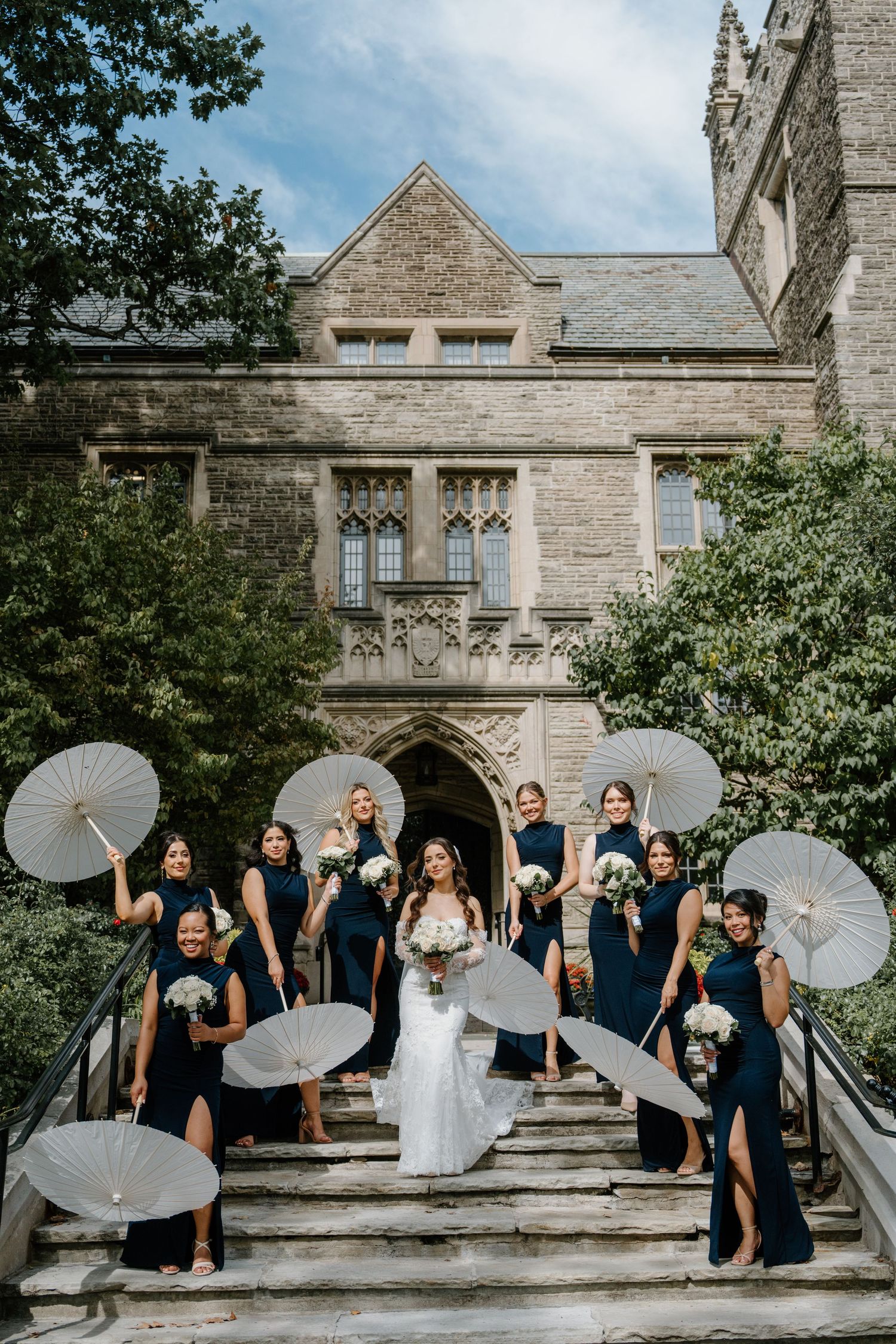 Wedding party poses with white parasols on stone steps of historic Gothic building in elegant outdoor setting.