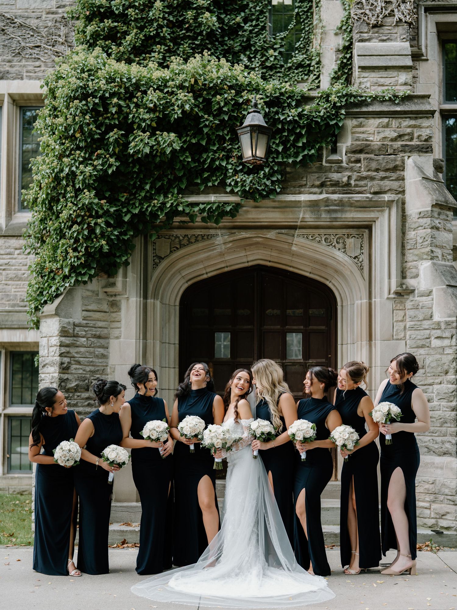 Wedding party in black dresses and white gown pose together against stone building in panoramic shot.