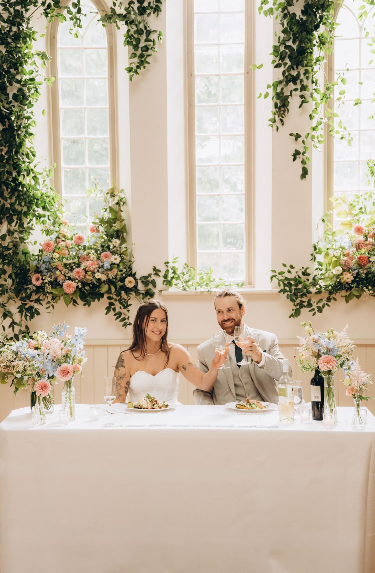 A wedding reception setup with floral decorations and greenery adorning tall windows in an elegant white room.