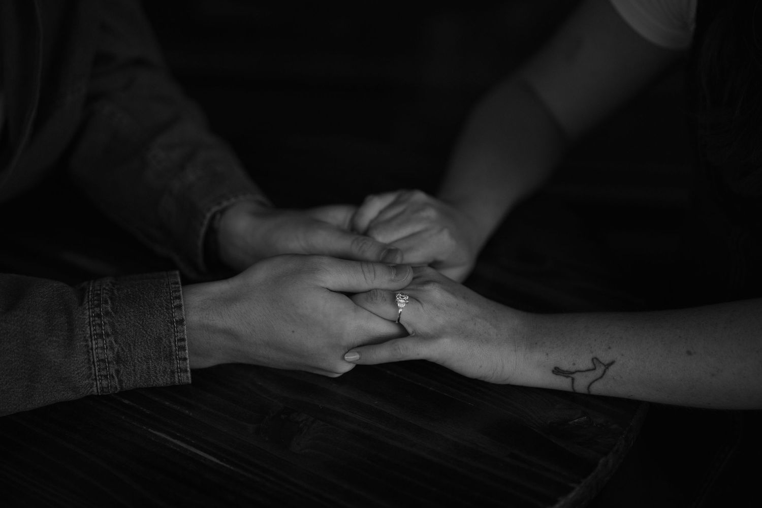 Two hands touching intimately with one having a small tattoo, captured in black and white lighting on dark background.