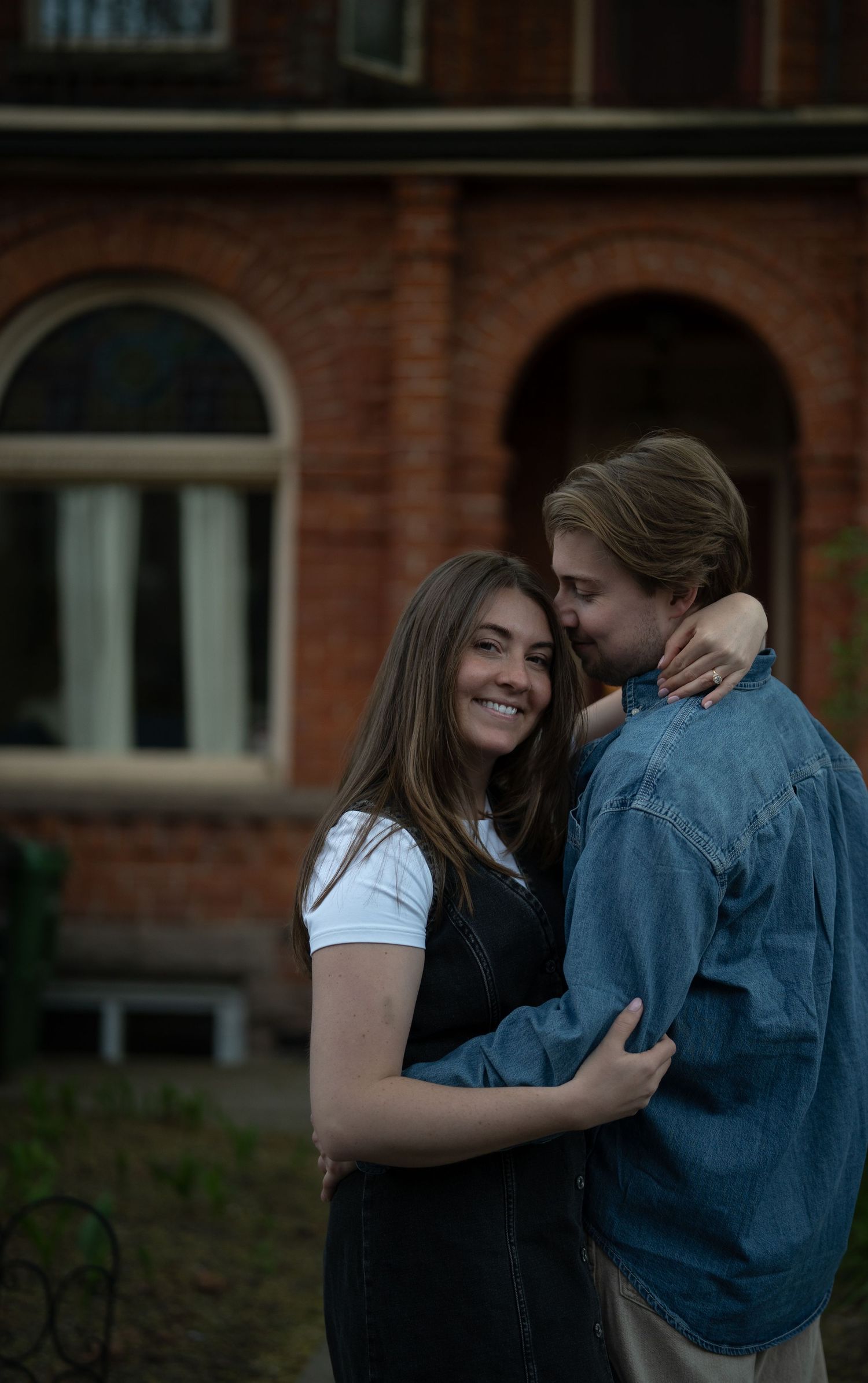 A romantic couple shares an intimate embrace while laughing together in front of a brick building with arched windows.
