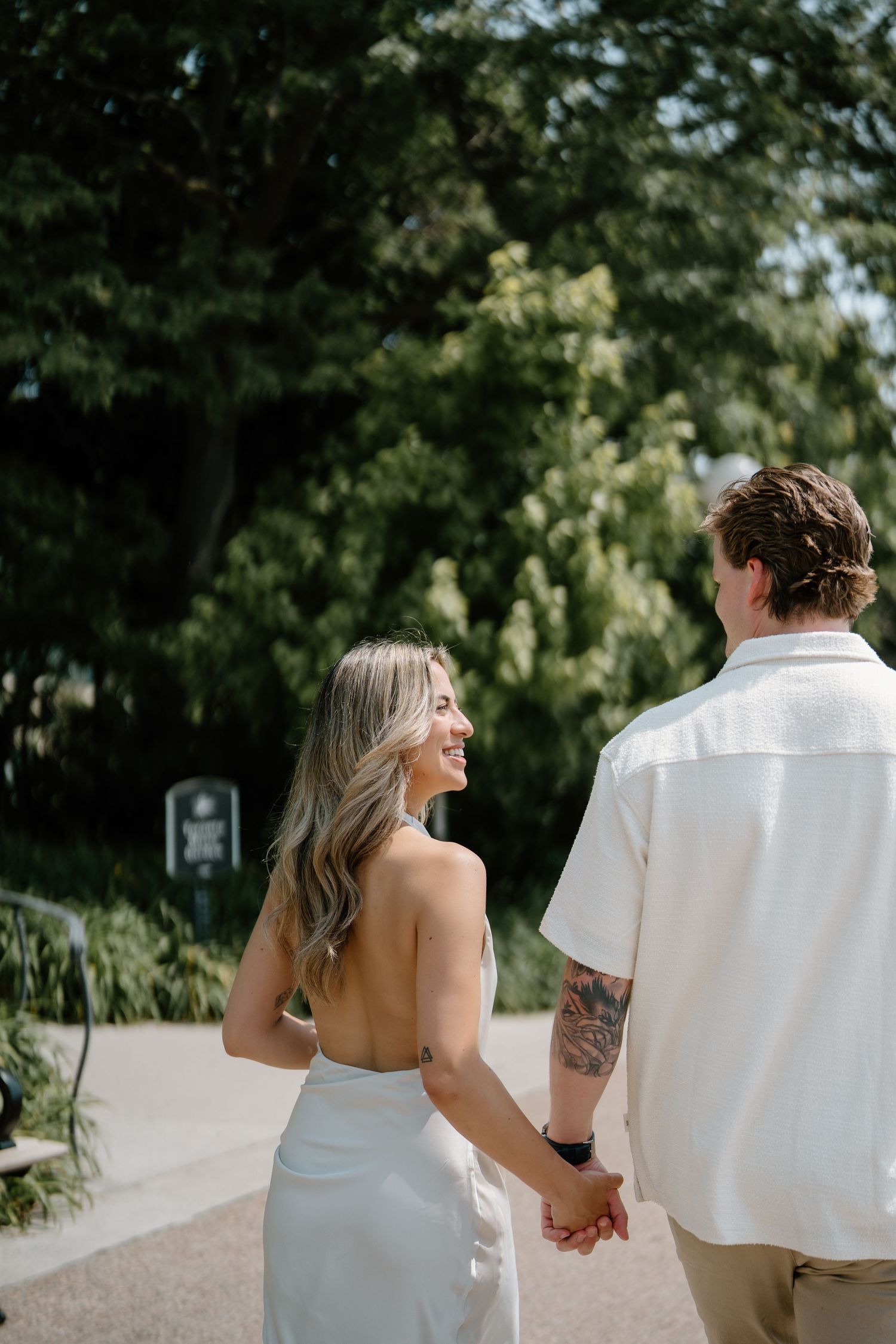 A couple holding hands and walking down a garden path, the bride in a white dress and groom in casual attire.