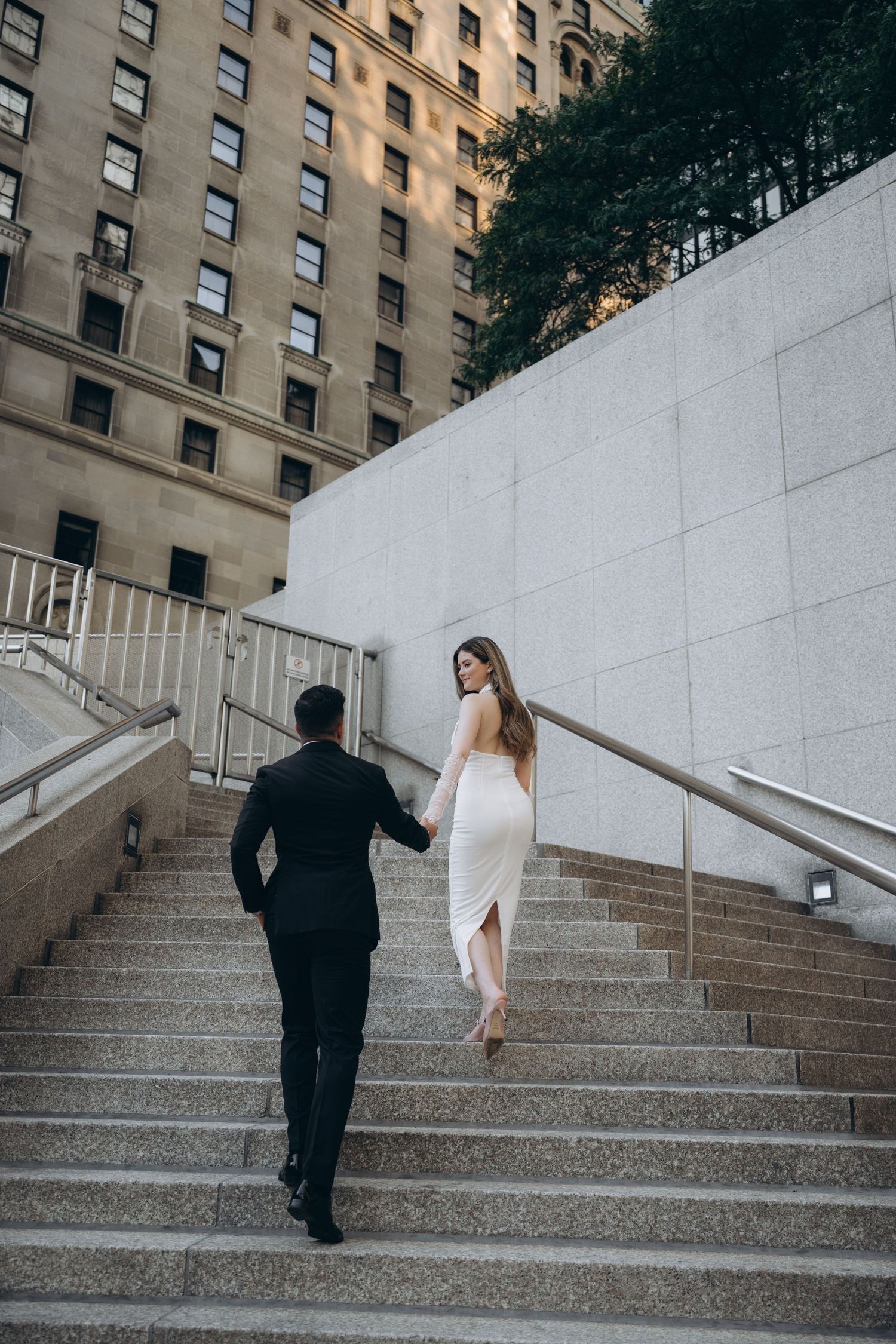 A couple ascends concrete stairs outside a tall apartment building, with the person in white leading the way up.
