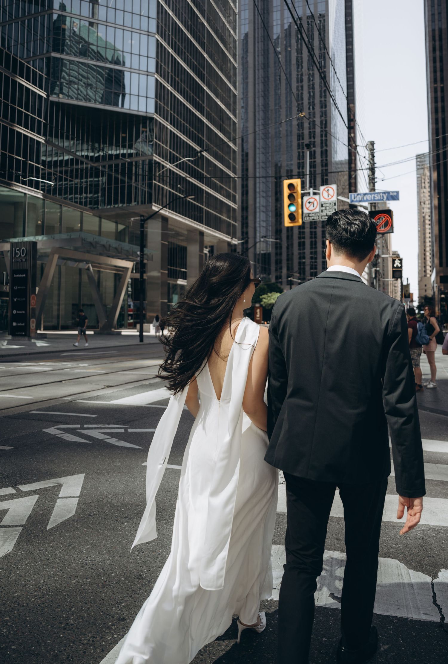 A wedding couple walks together on a downtown street corner surrounded by modern glass office buildings.