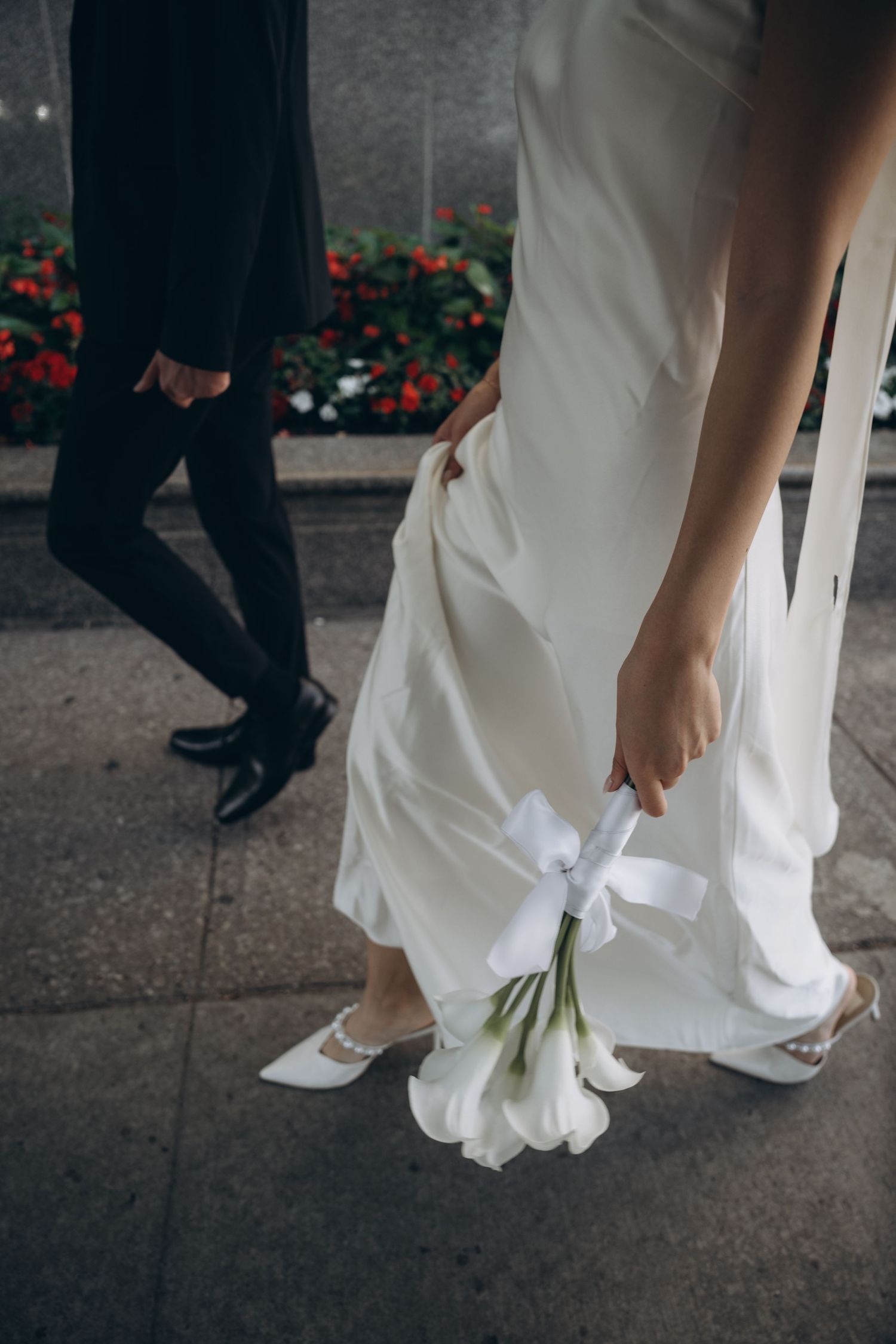 A close-up view of elegant white wedding shoes and dress hem while walking on concrete pavement.