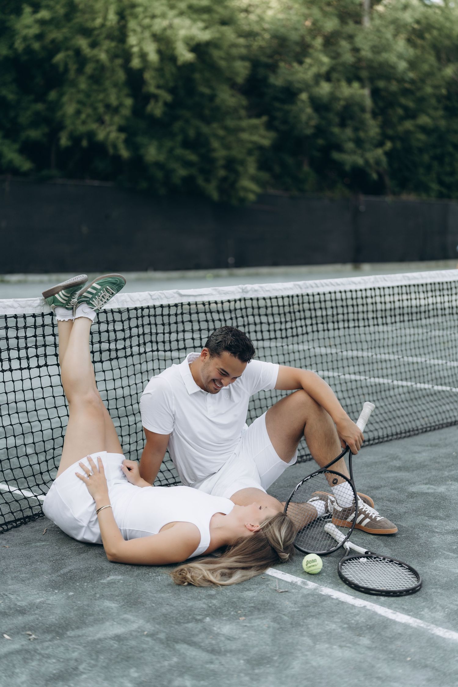 A couple in white tennis attire laying on a tennis court next to rackets and balls after playing a match.
