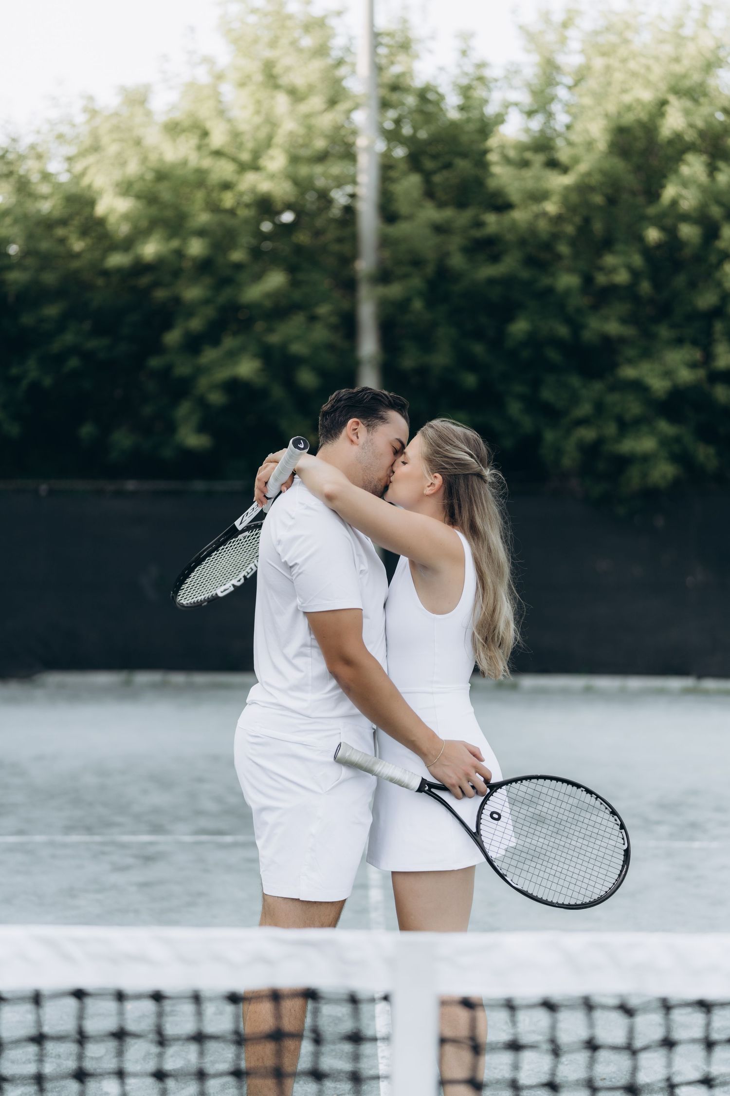 A couple in white athletic wear share a romantic moment on a tennis court surrounded by trees.