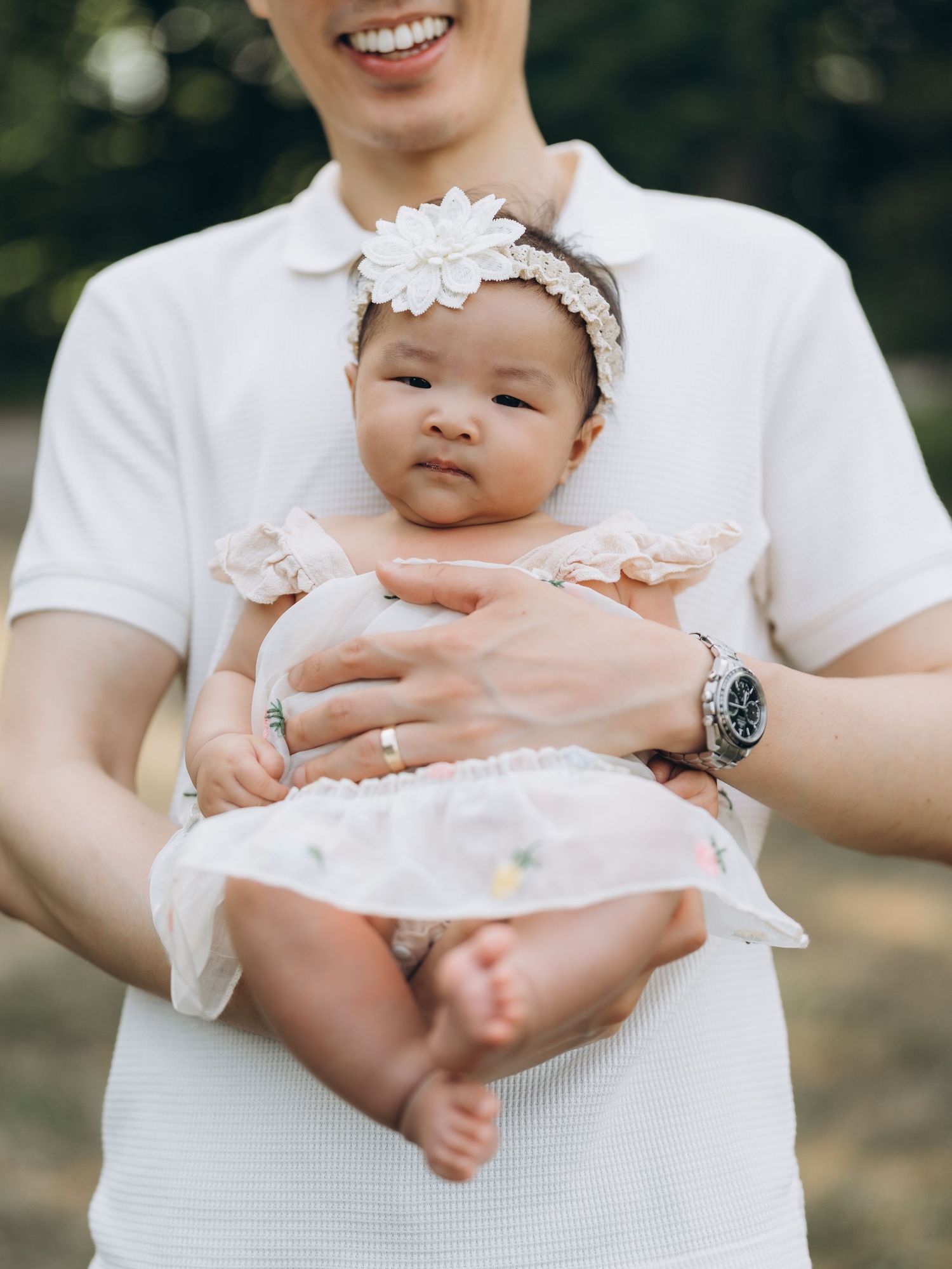 A baby wearing a white lace headband is held against someone in a white shirt.