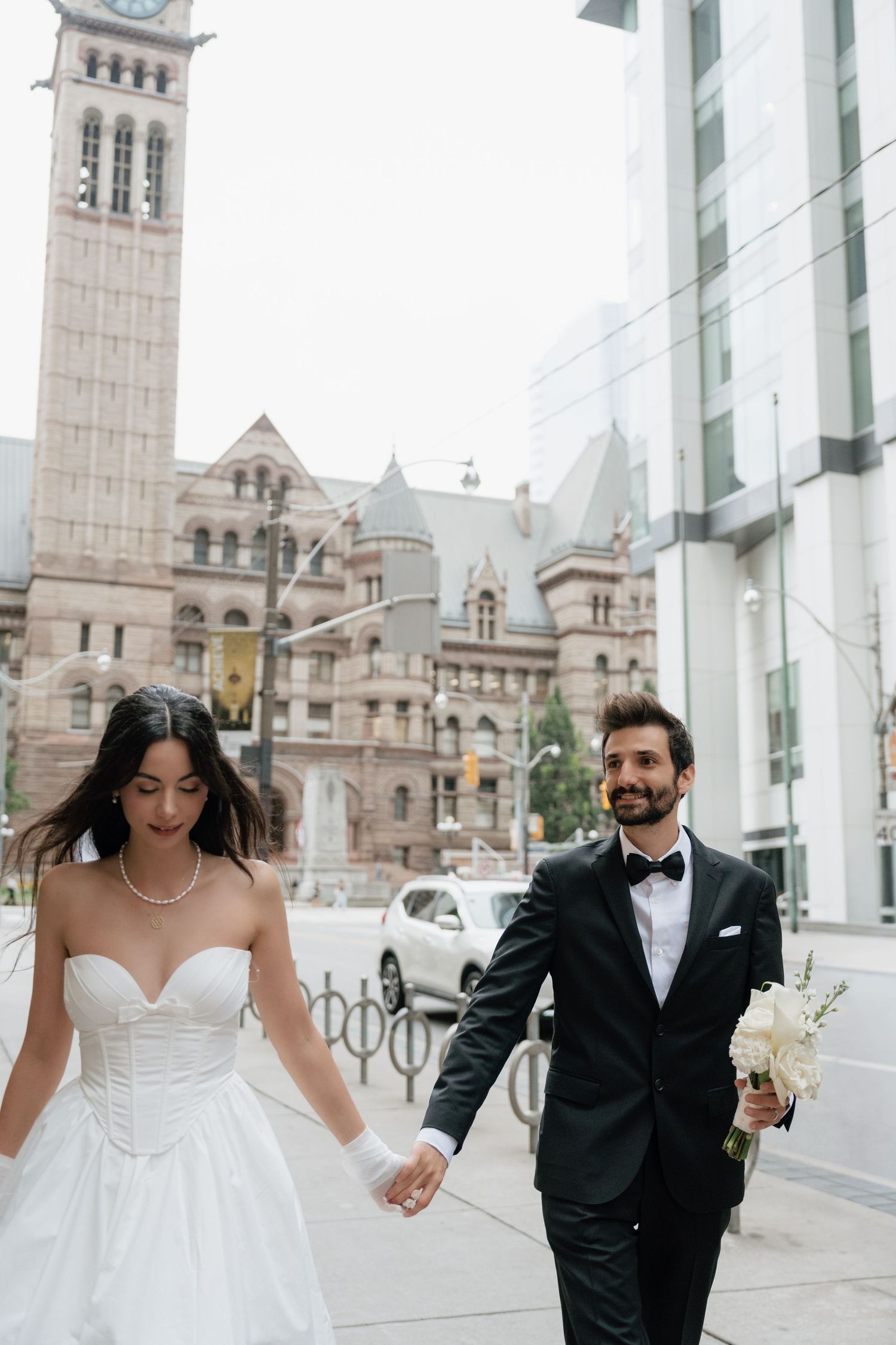 A wedding couple runs joyfully through a modern architectural space with white columns and a historic building in the background.