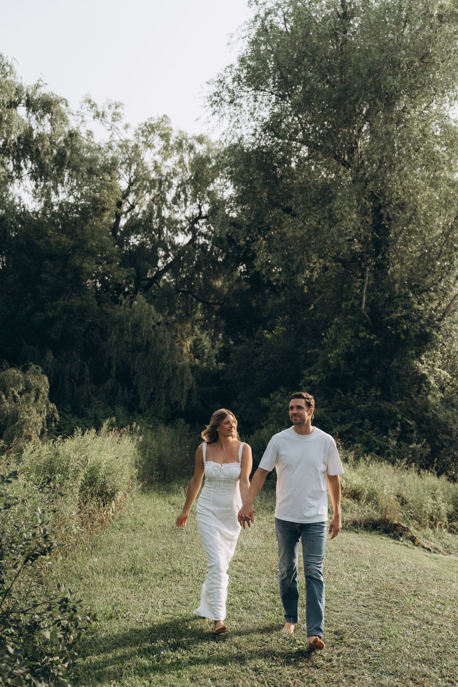 A couple walks hand in hand up a grassy hill surrounded by trees on a sunny day.