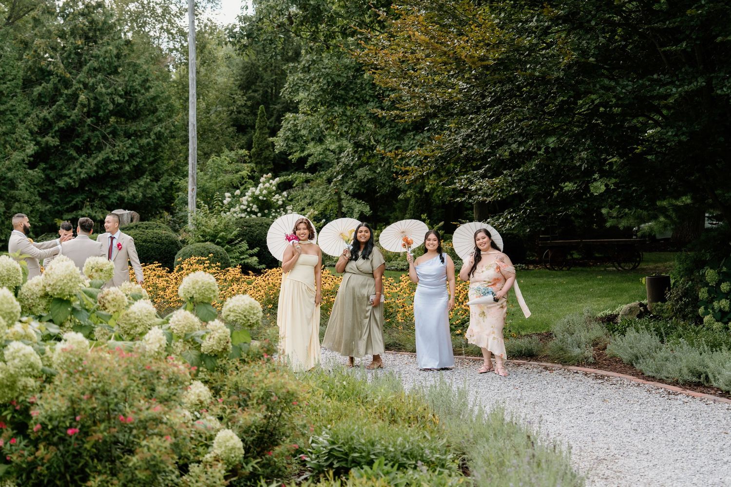 A wedding party walks along a gravel path surrounded by lush gardens and greenery on a summer day.