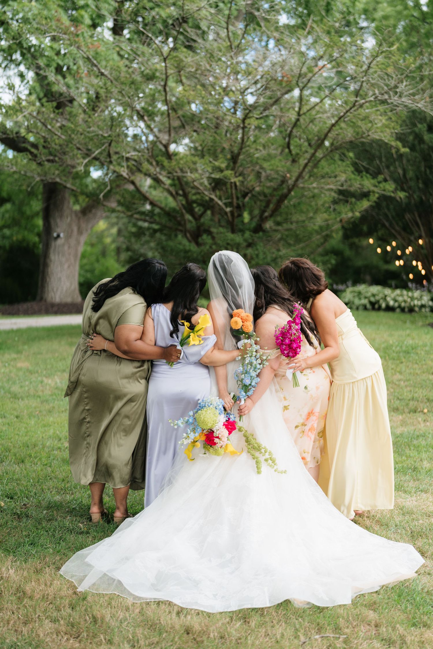 A group of bridesmaids in colorful dresses pose together with the bride in a white gown on a grassy lawn under trees.