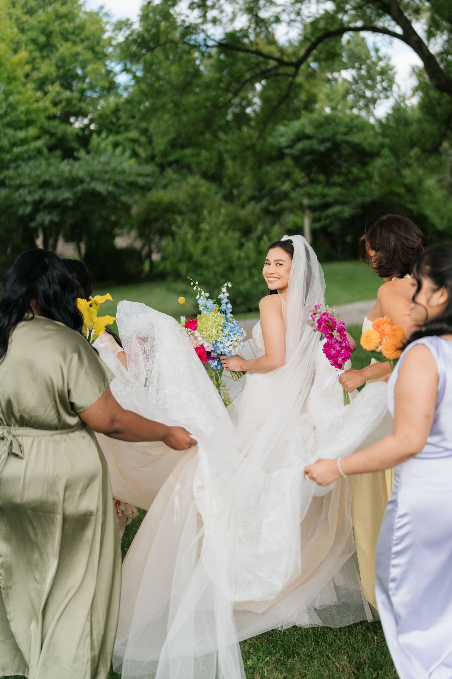 A wedding party in a line holds up the bride's flowing white dress train while walking through a lush green garden setting.