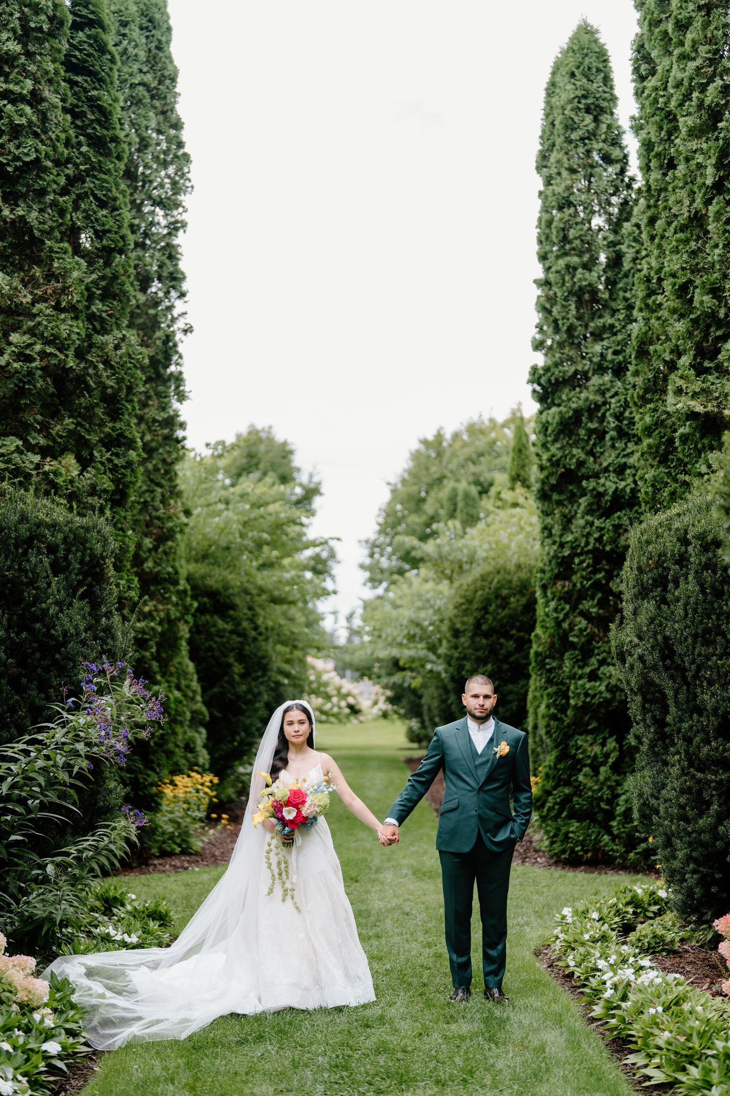 A couple in wedding attire walks through a lush garden path lined with tall cypress trees and colorful flowers.