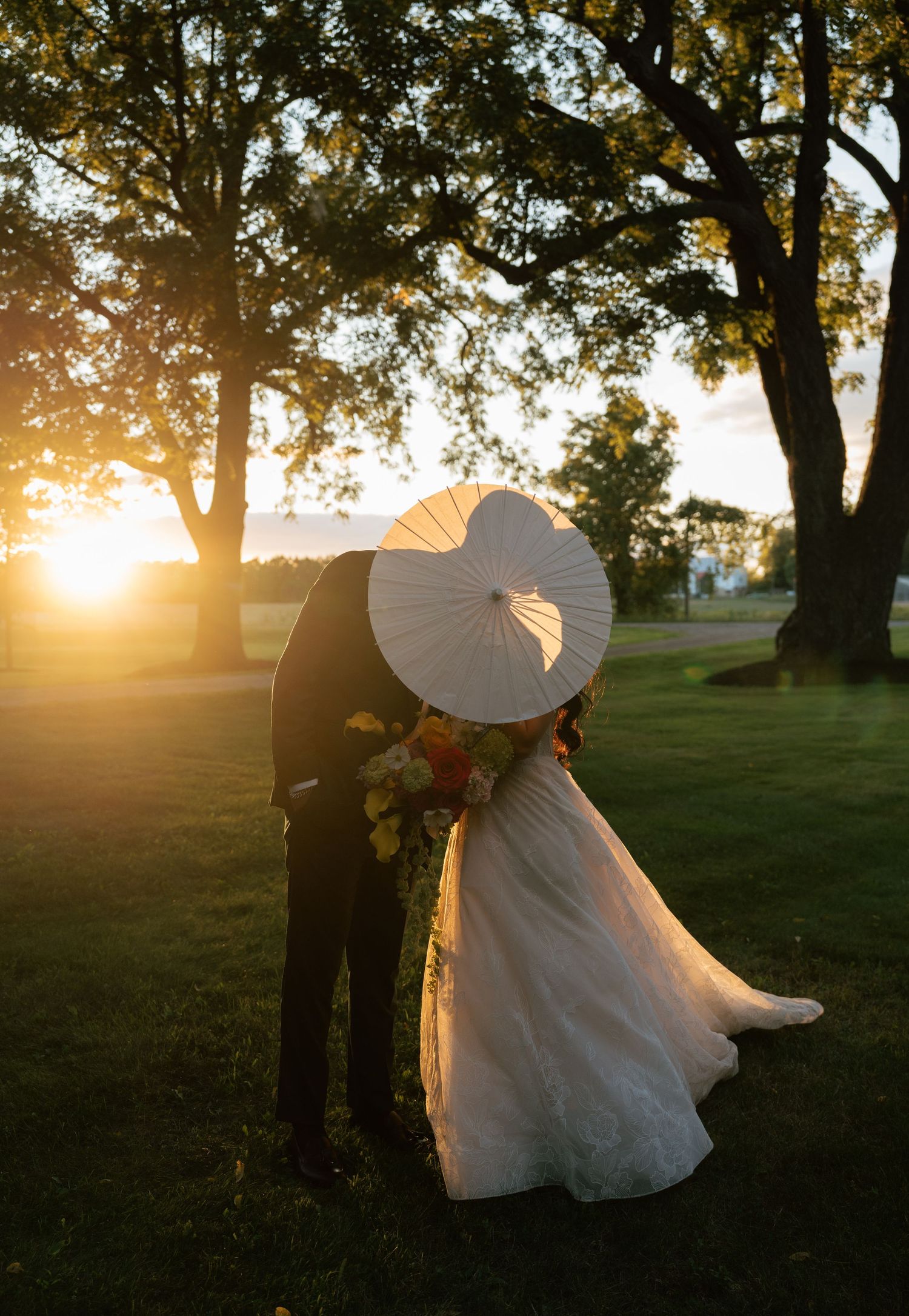 Silhouetted wedding couple poses against a golden sunset with trees creating a dreamy outdoor backdrop.