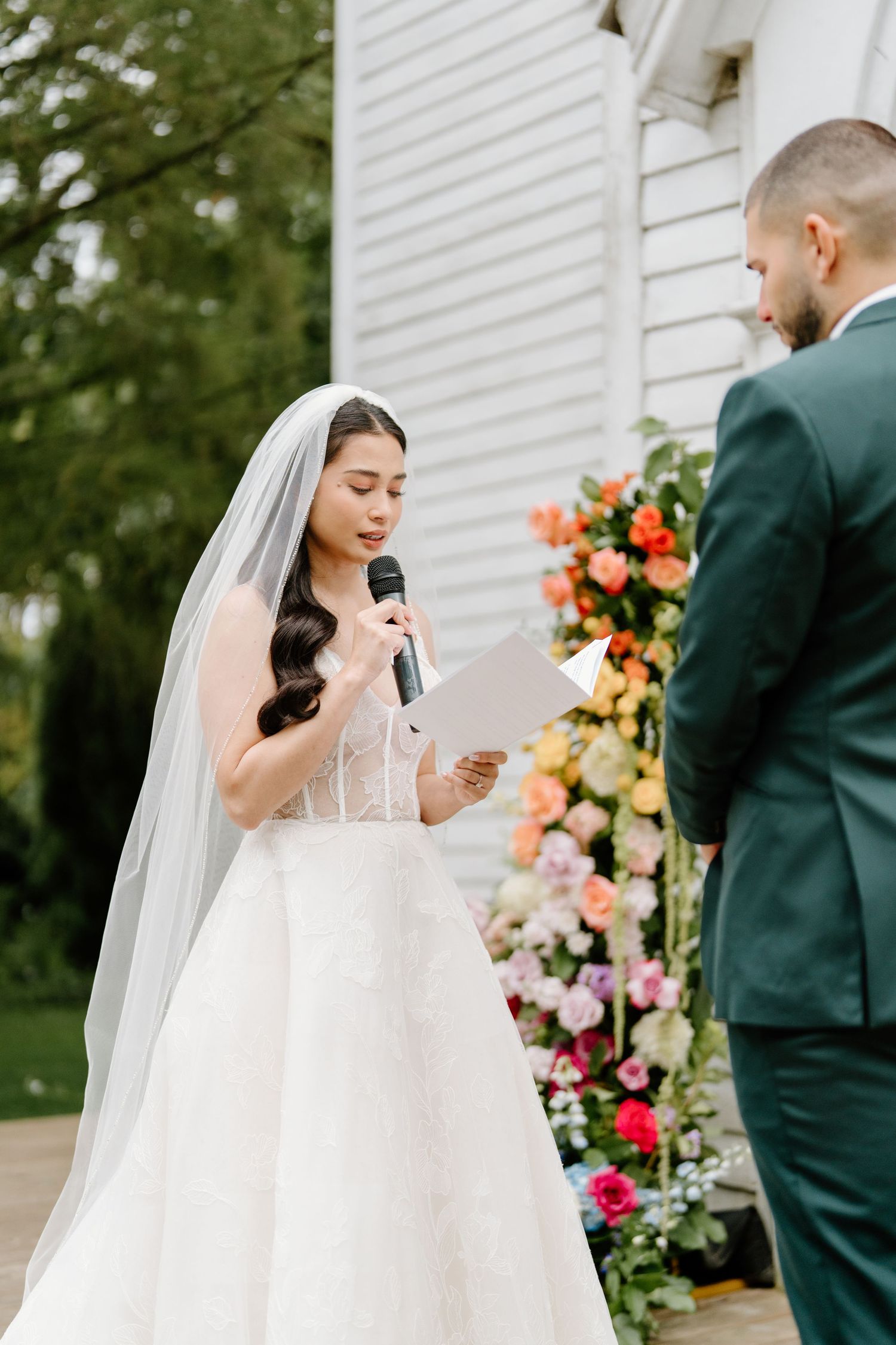 A bride in a white gown and veil reacts emotionally during an outdoor wedding ceremony with colorful floral arrangements.
