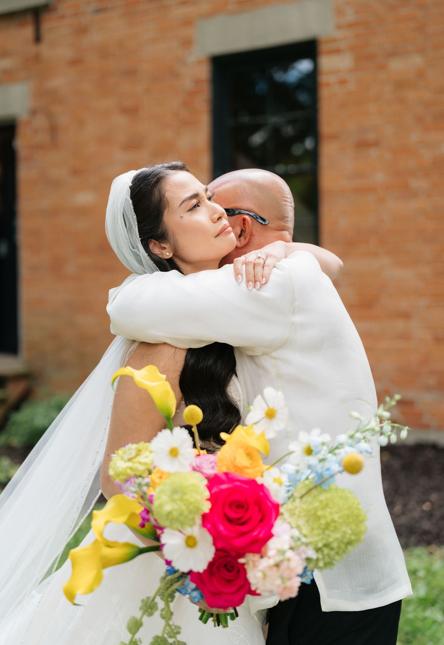A romantic wedding couple embraces in front of a brick building with colorful spring flowers in the foreground.