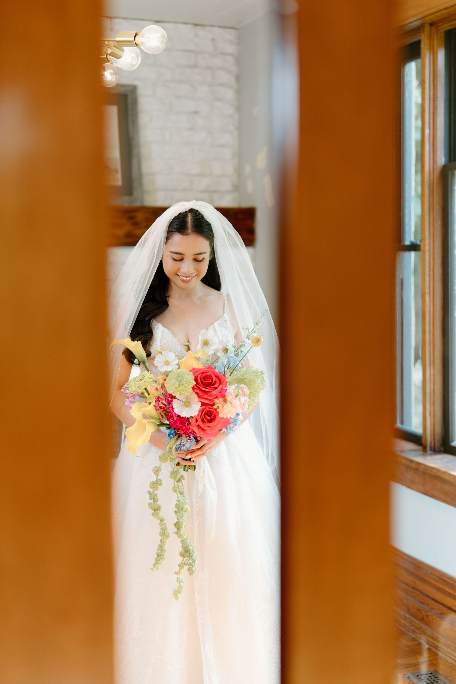 A bride in a white gown and veil holds a vibrant bouquet of red and yellow flowers in a sunlit indoor setting.