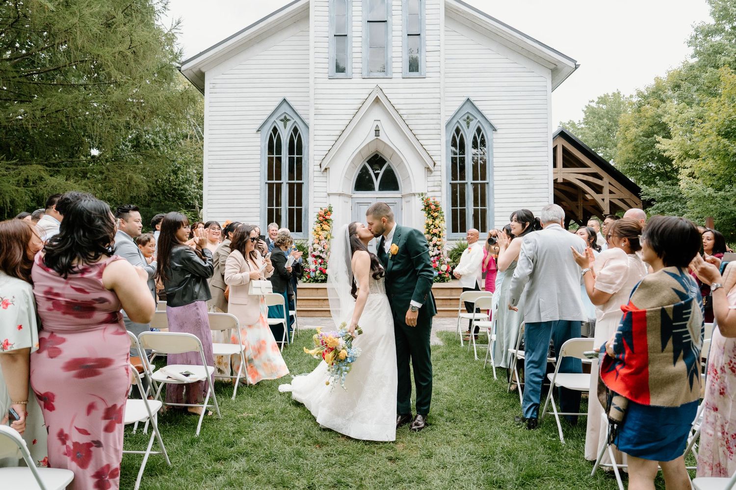 A wedding ceremony takes place outside a charming white chapel with Gothic windows and guests seated on folding chairs.