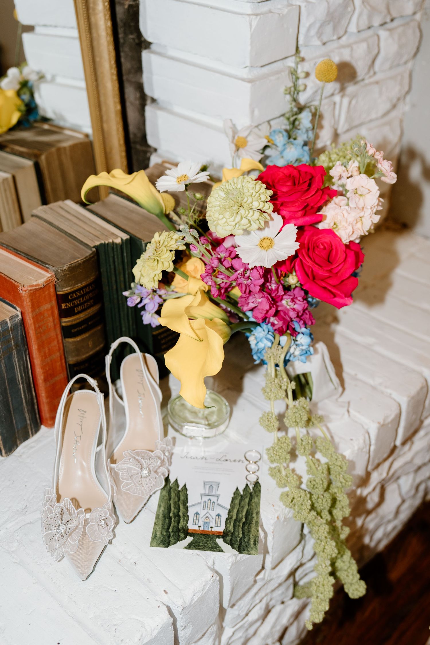 Elegant bridal shoes displayed with vintage books and colorful floral arrangements on a white mantel.
