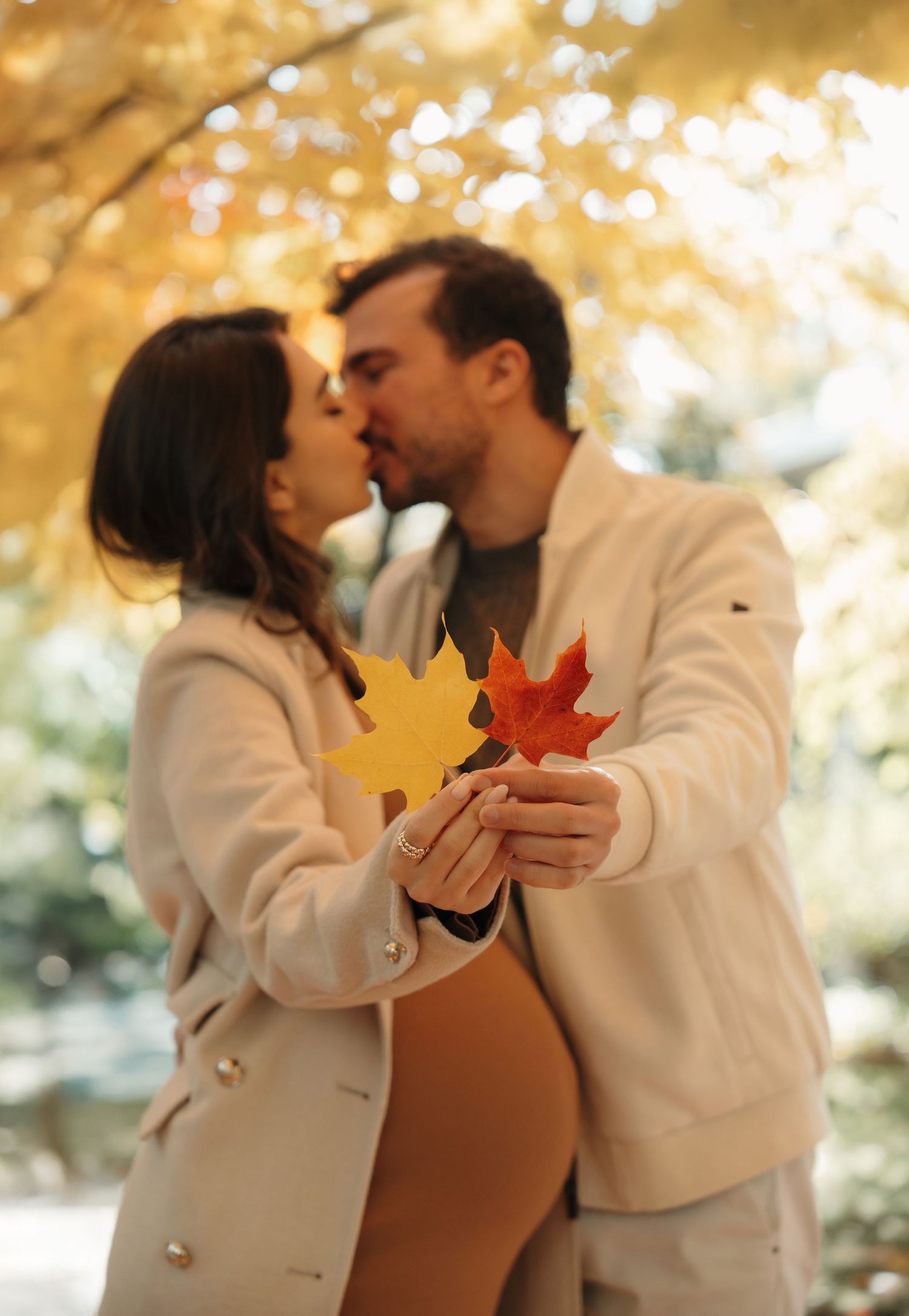 A romantic autumn moment with two people holding colorful maple leaves while sharing a tender embrace in beige coats.