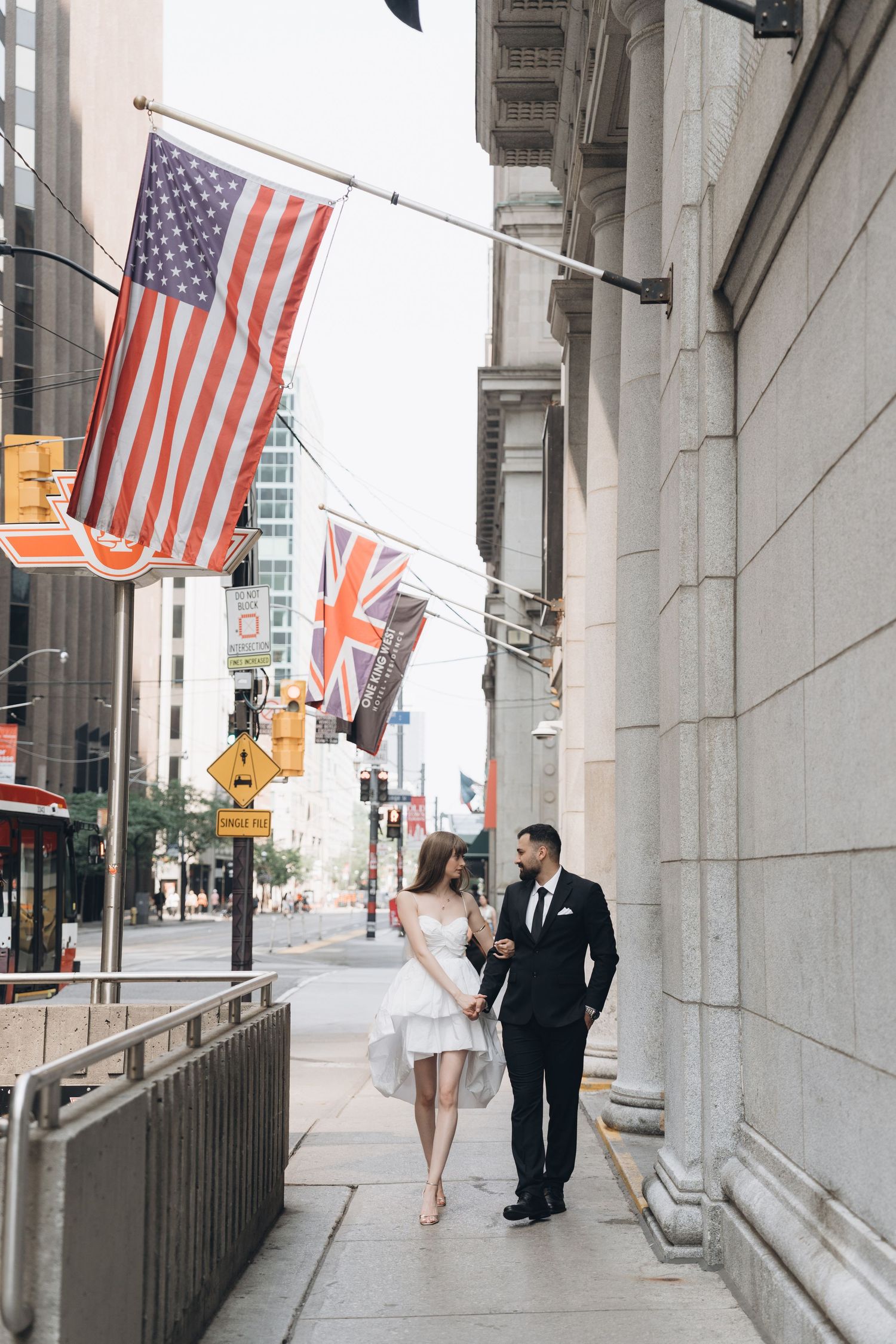 Couple walking along urban sidewalk under American and British flags displayed on a city building exterior.