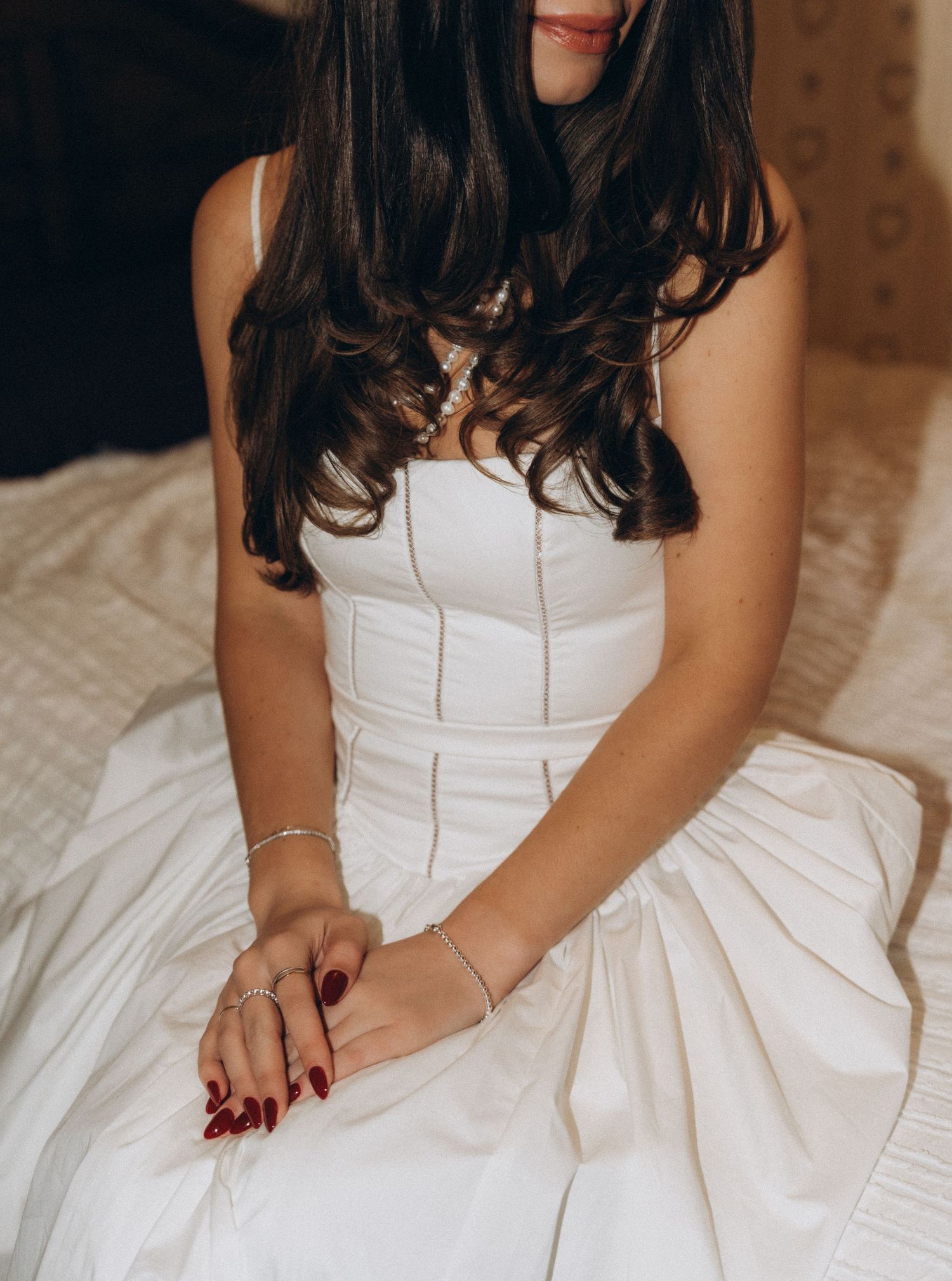 Woman wearing a white wedding dress sits elegantly on a bed, showcasing delicate details and feminine pose.