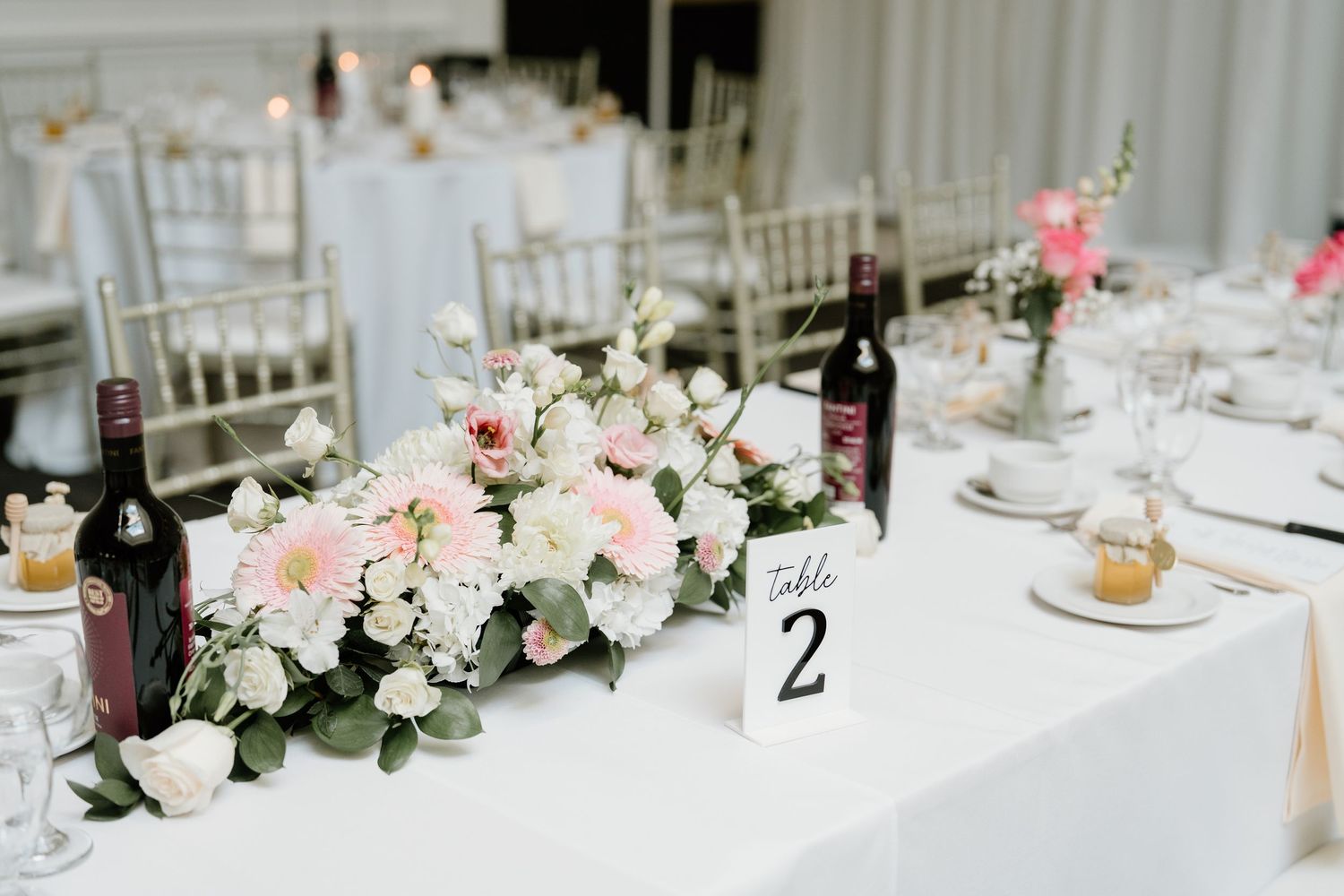 Long banquet table decorated with pink and white roses, greenery garlands and wine bottles for a wedding reception.