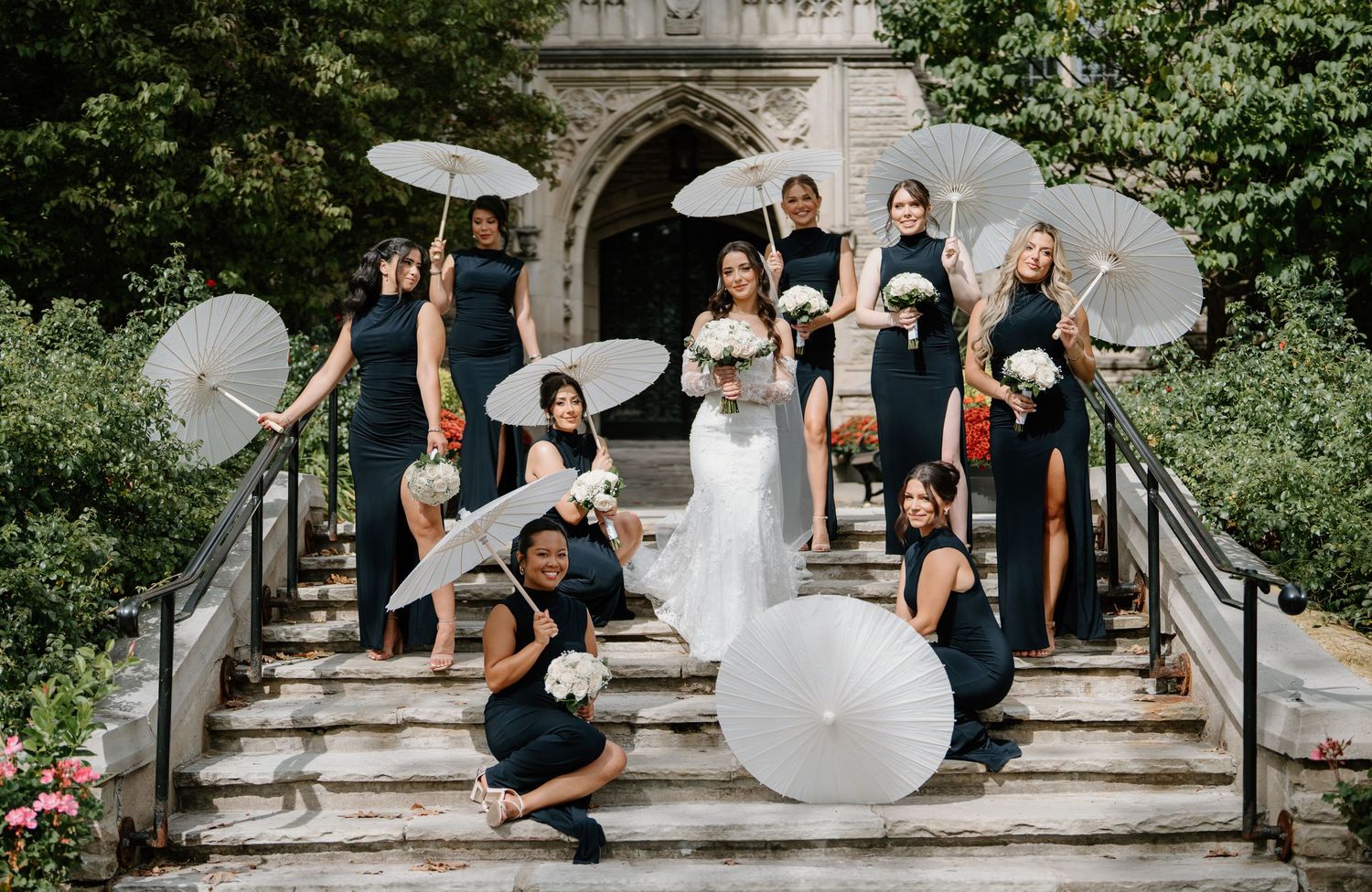 Wedding party poses playfully on stone steps with white umbrellas in an outdoor garden setting.