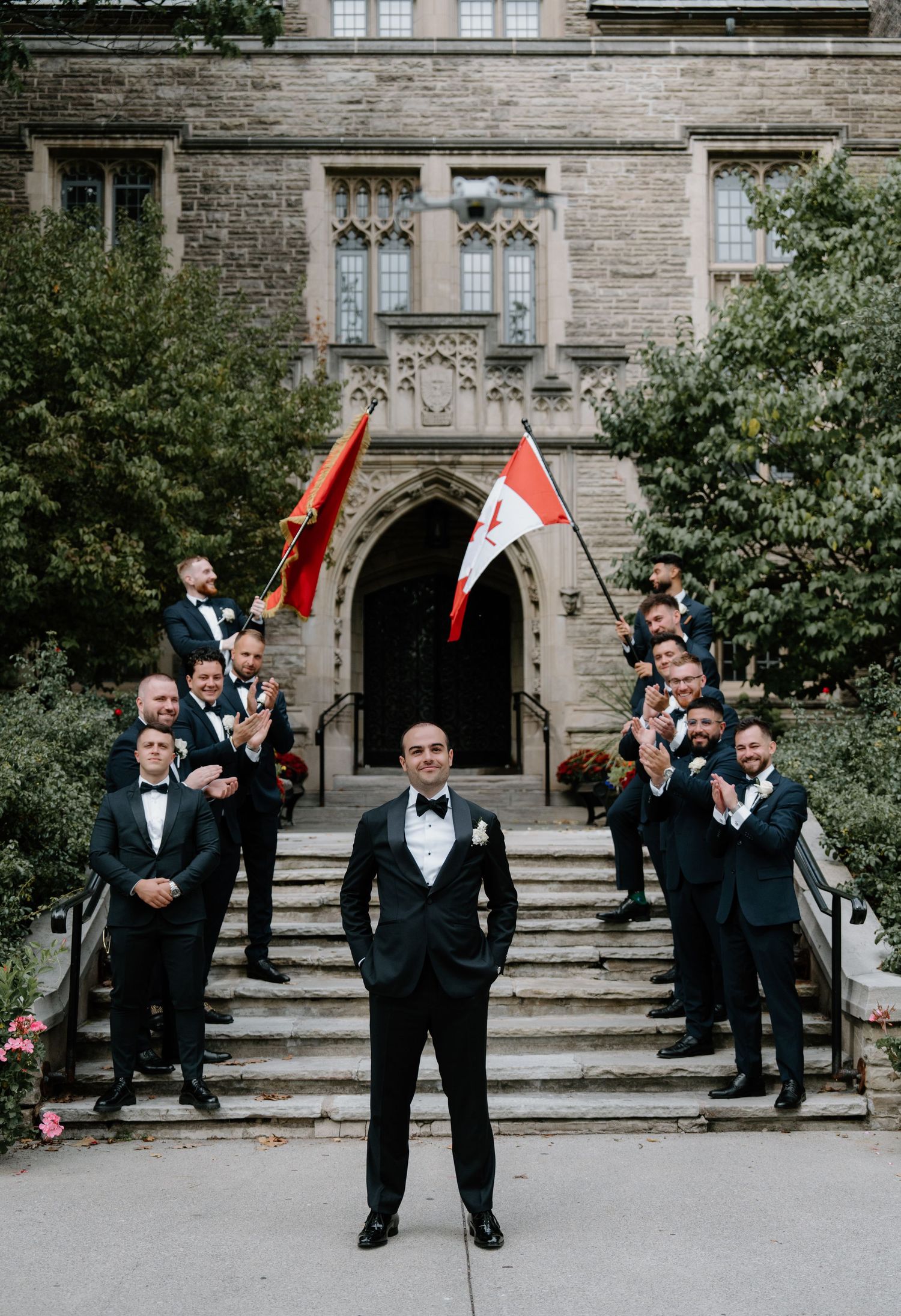 Groomsmen in black tuxedos pose for creative sequence of photos with red flags on stone building steps.