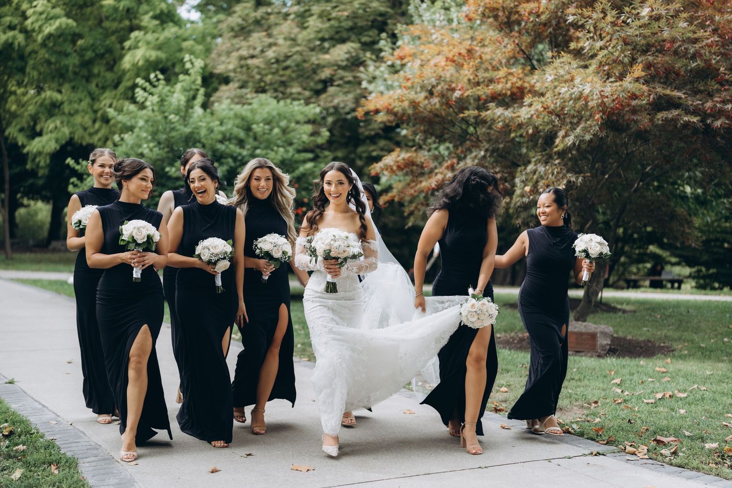 Wedding party in black attire poses playfully while carrying bride in white dress through garden setting.