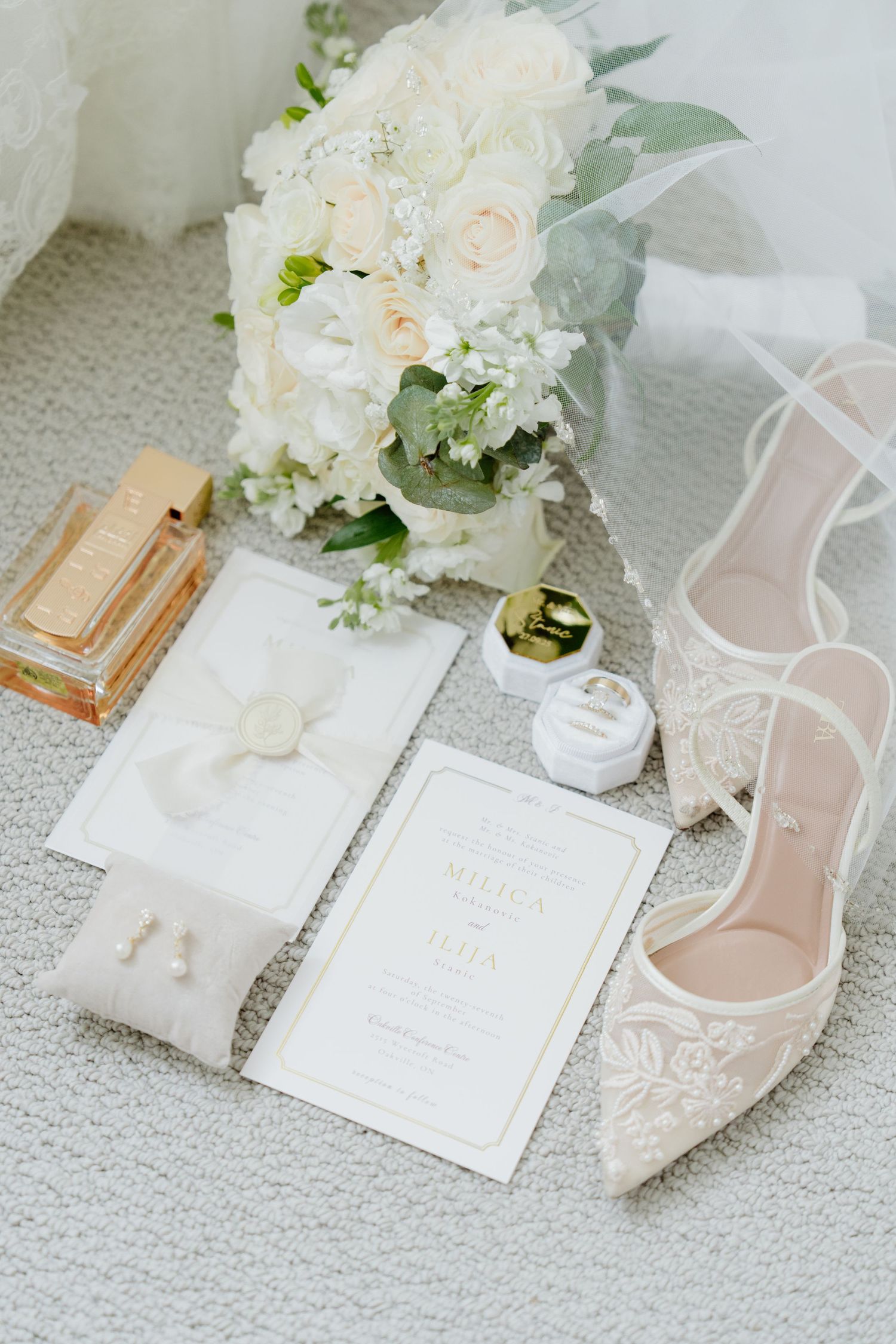 Wedding accessories including white floral bouquet, invitation cards, delicate shoes and rings arranged on light carpet.