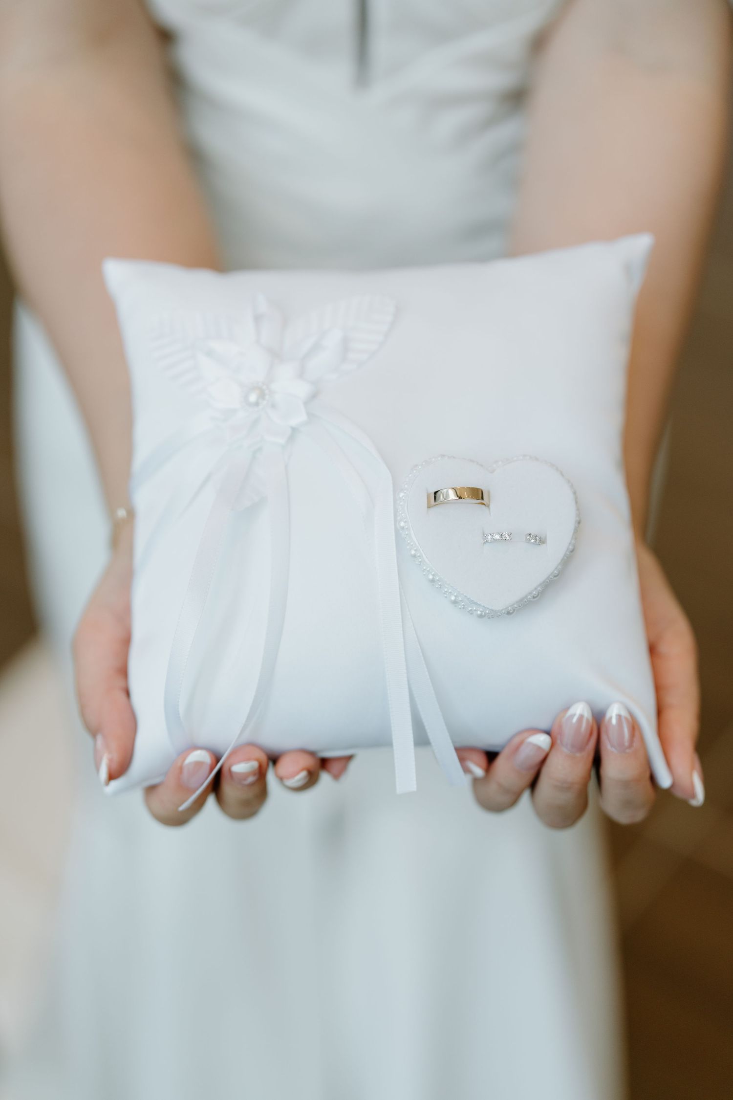 A white wedding ring pillow with decorative bow and small heart detail held by hands with manicured nails.