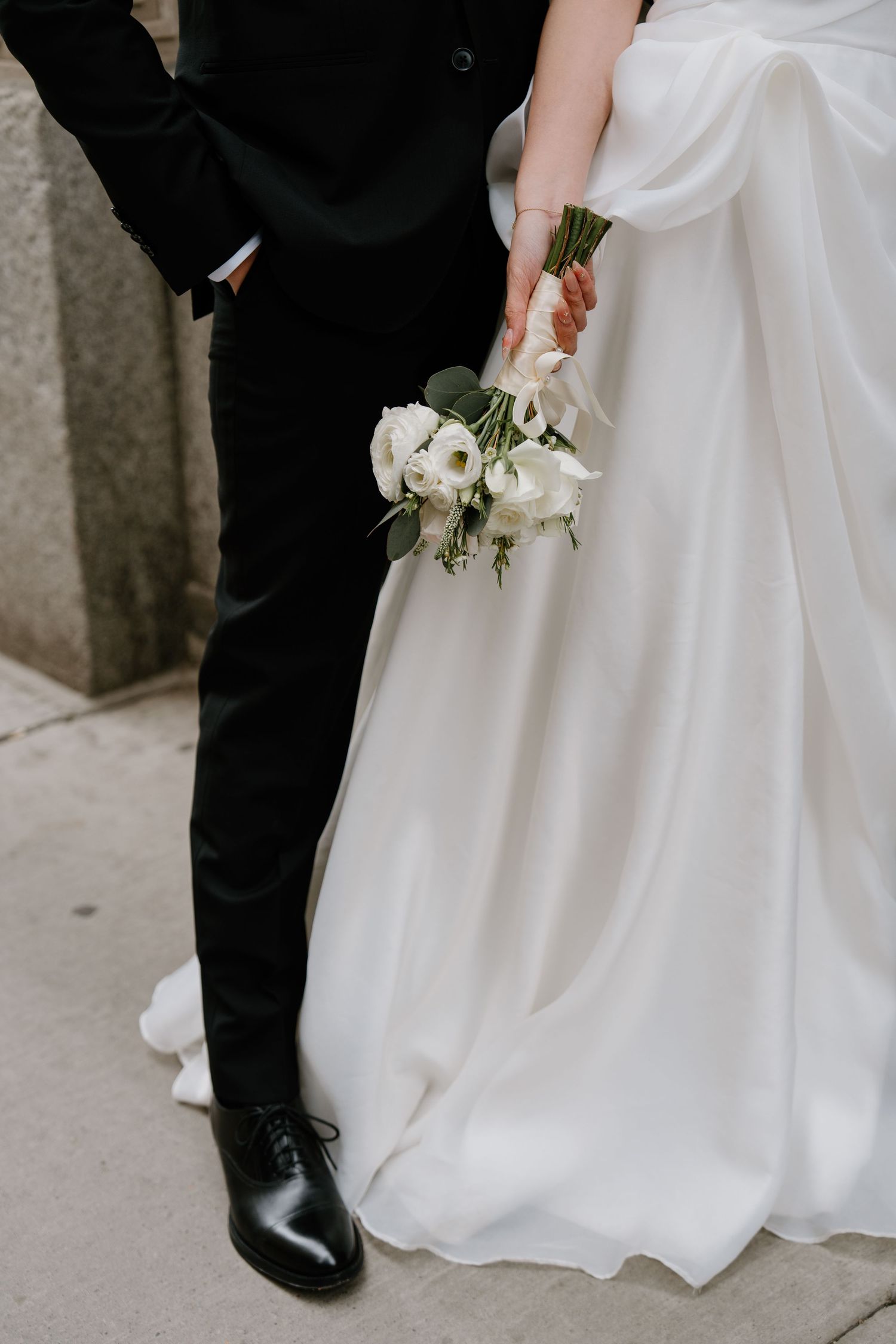 Close-up view of bride in white gown holding white floral bouquet standing next to groom in black suit and dress shoes.