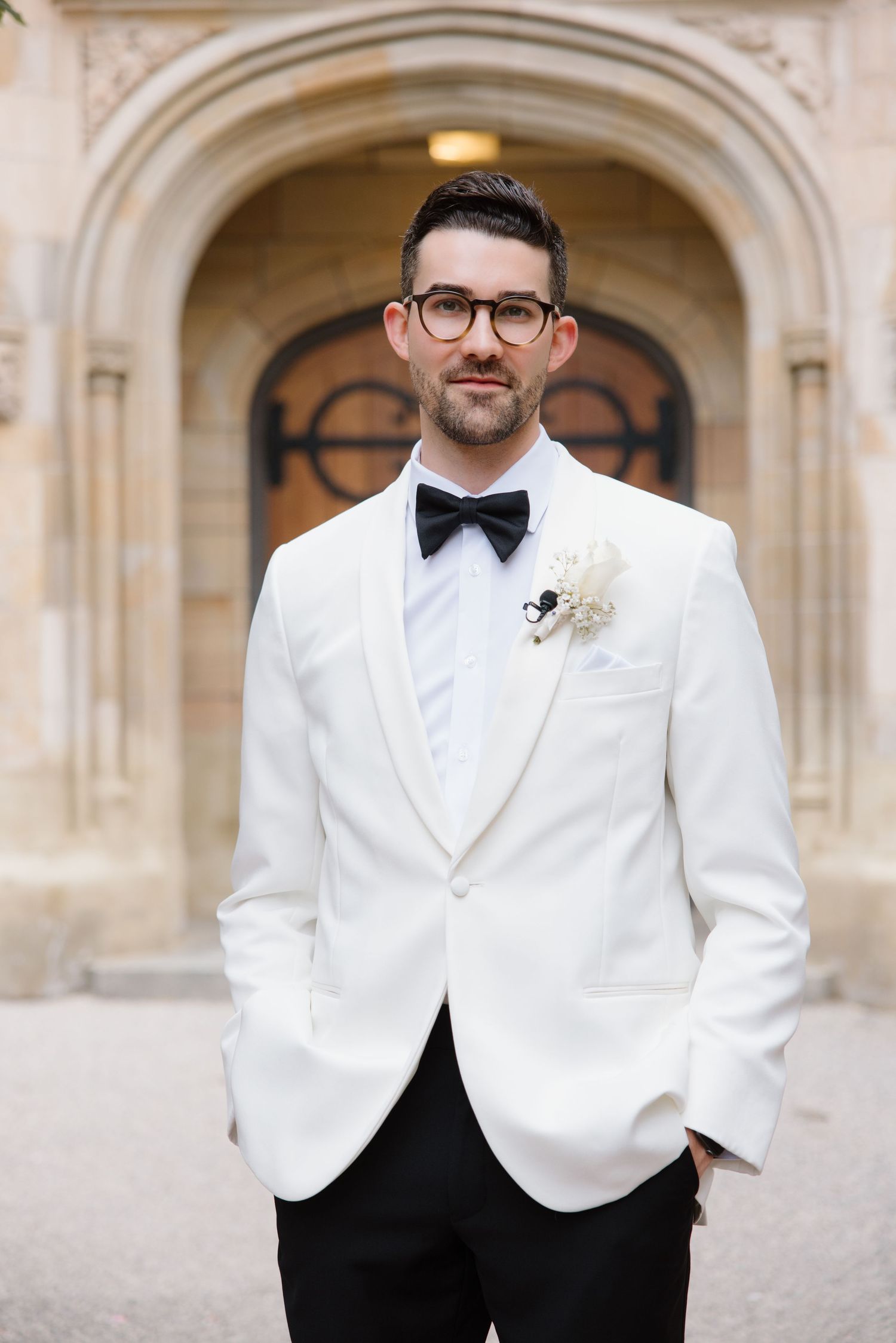 Groom in classic white dinner jacket and black bow tie standing in elegant stone archway setting.