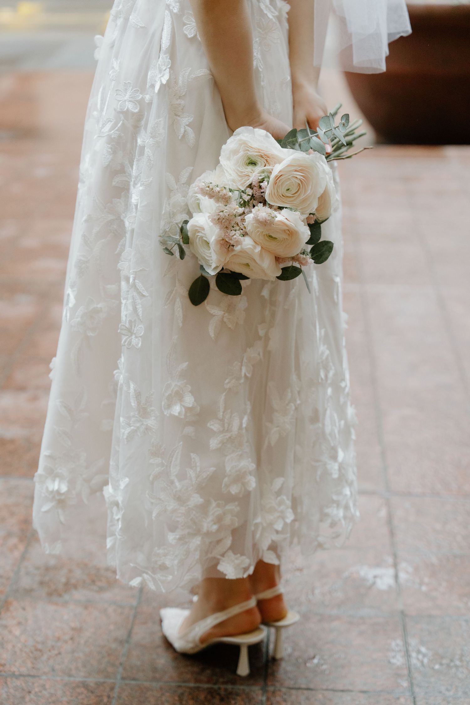 A delicate white wedding bouquet with roses and greenery held against a lace wedding dress and white heels.