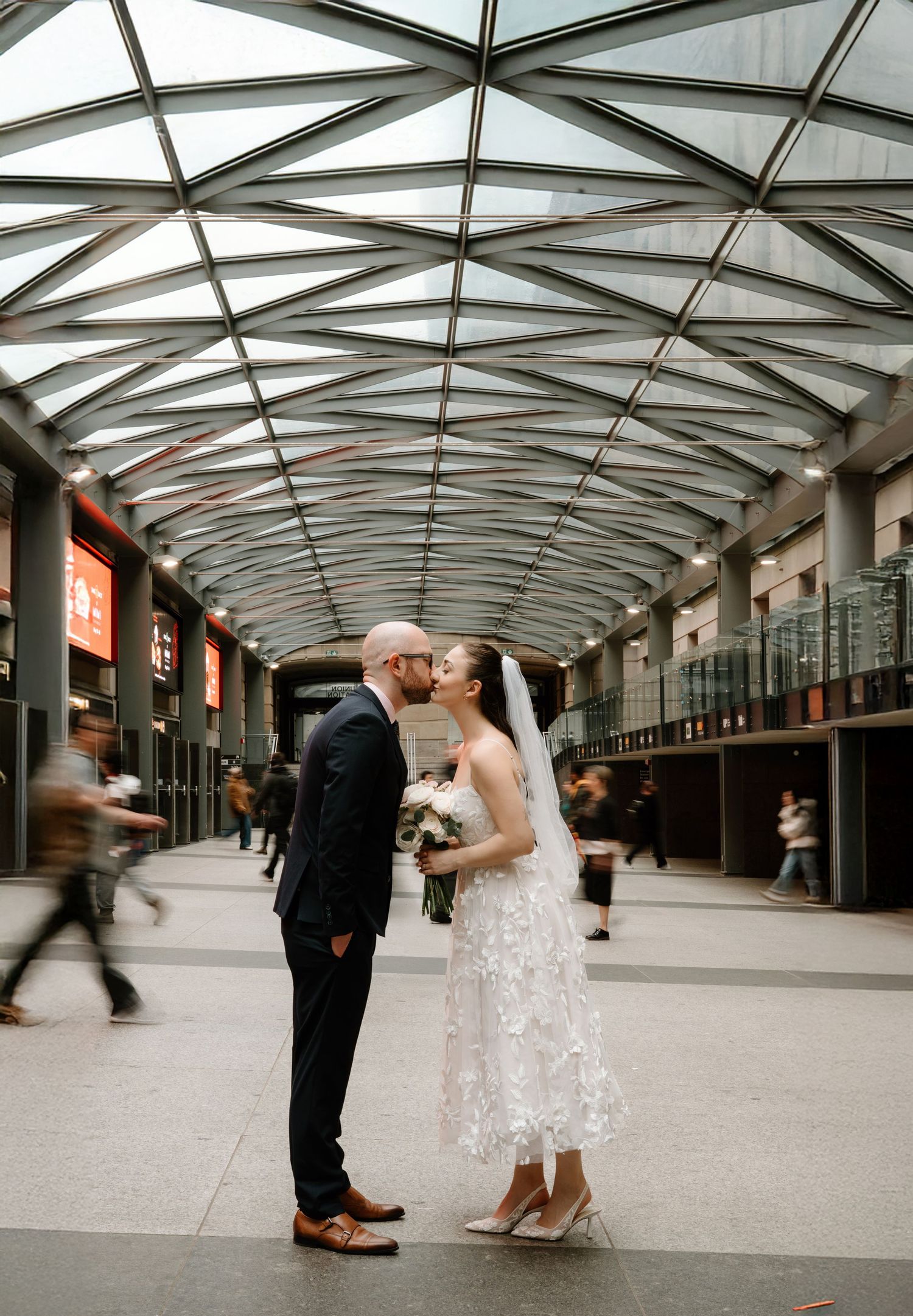 Romantic wedding couple dancing in a modern glass-covered walkway with architectural leading lines.