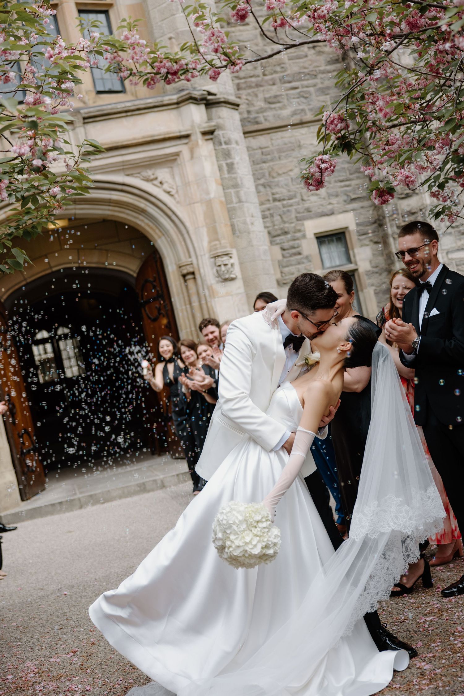 Newlyweds share romantic kiss under stone archway as guests shower them with confetti at church wedding celebration.