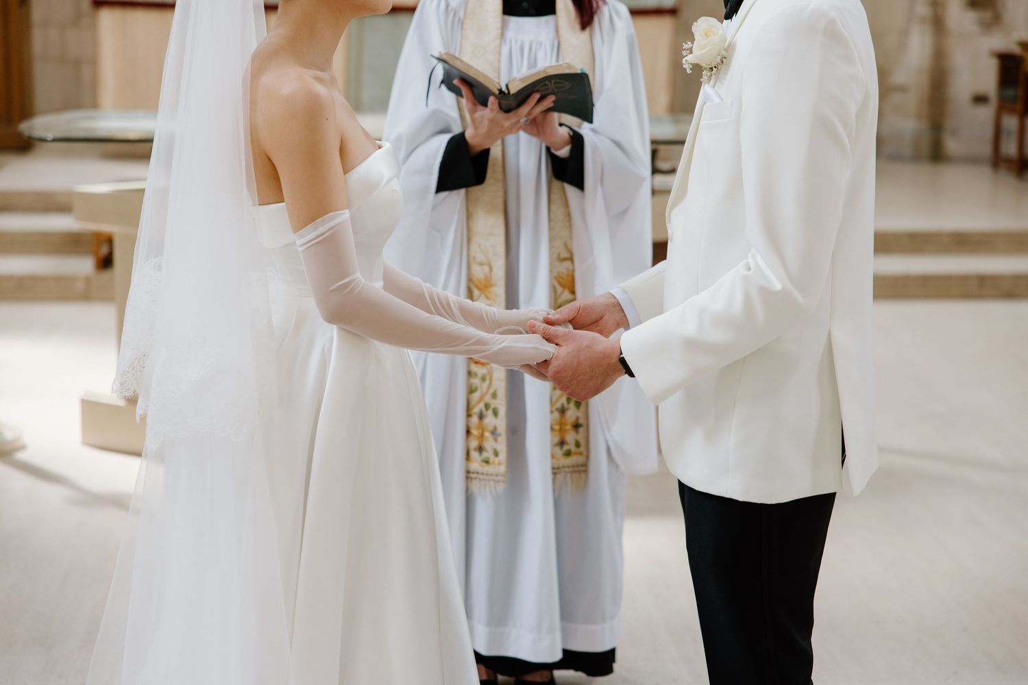 A couple holds hands during their wedding ceremony as an officiant in white robes presides over their intimate indoor service.