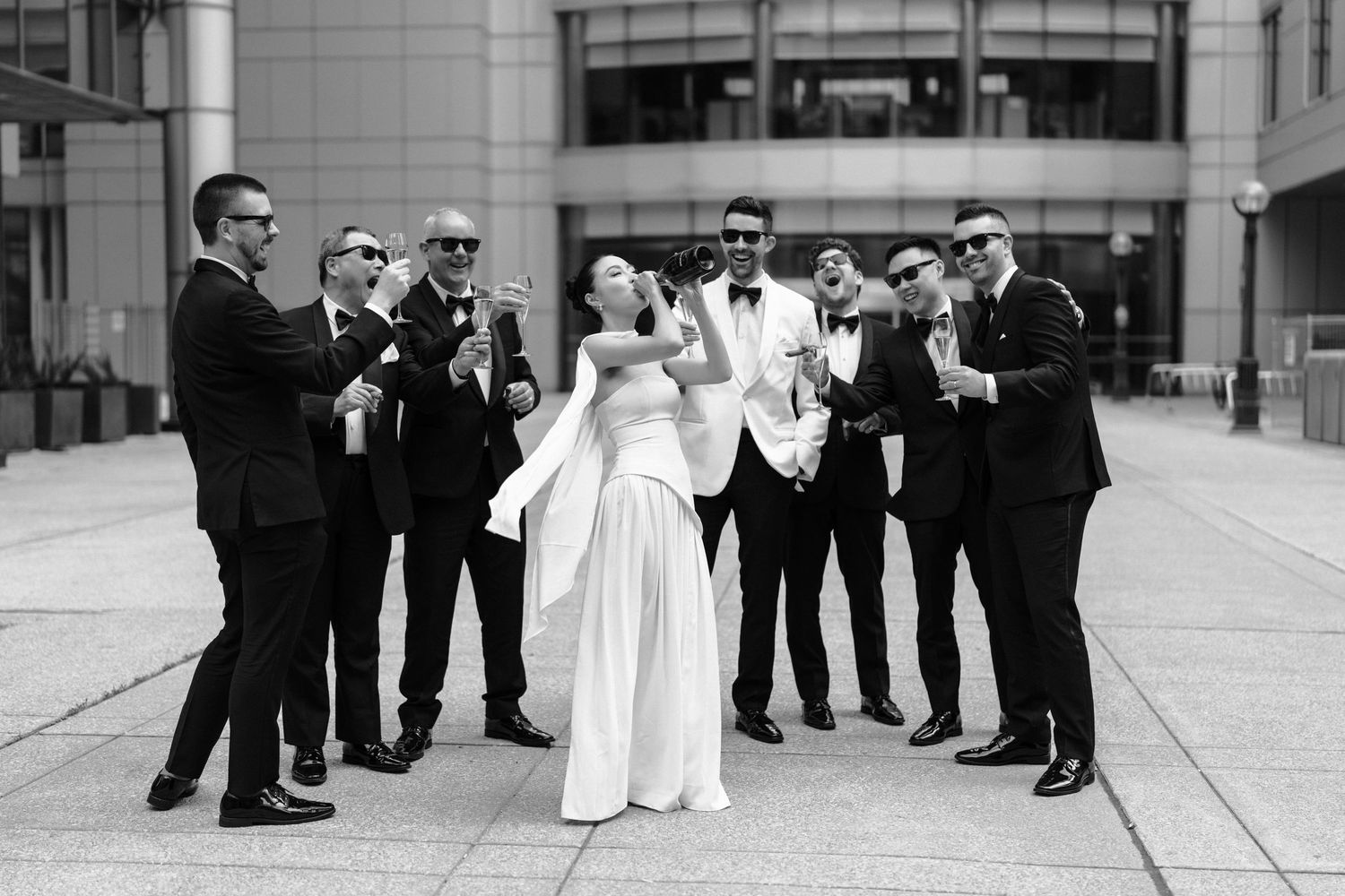 A wedding party in formal attire celebrates together on a modern outdoor plaza in black and white.