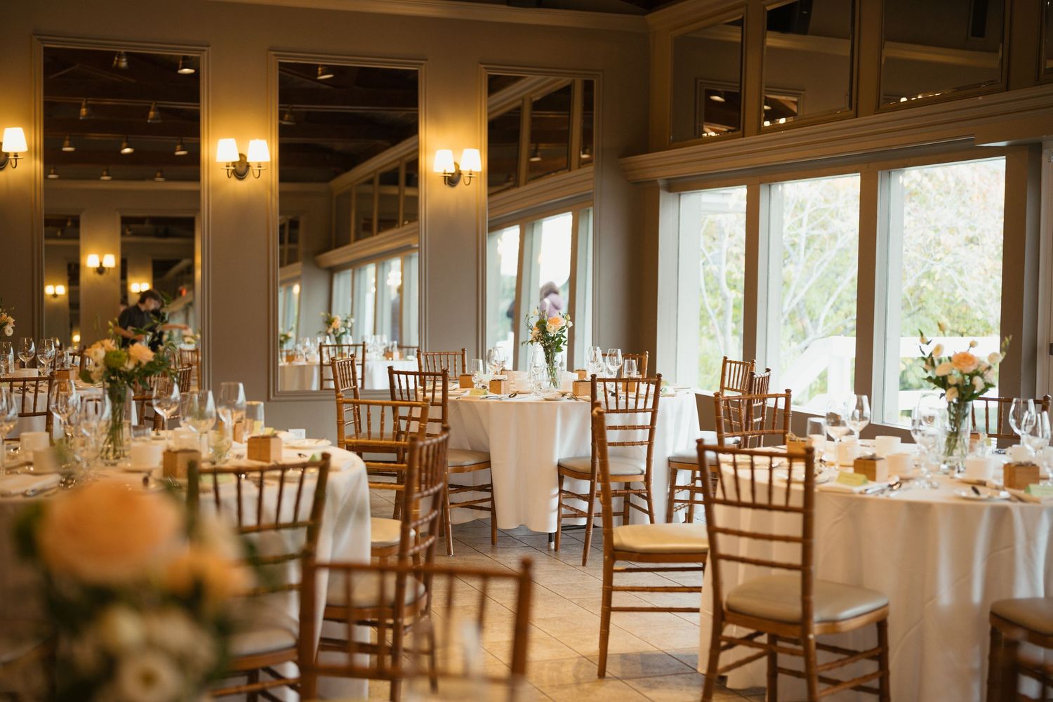 Light-filled wedding venue with classic round tables, floral centerpieces, and wooden chairs.