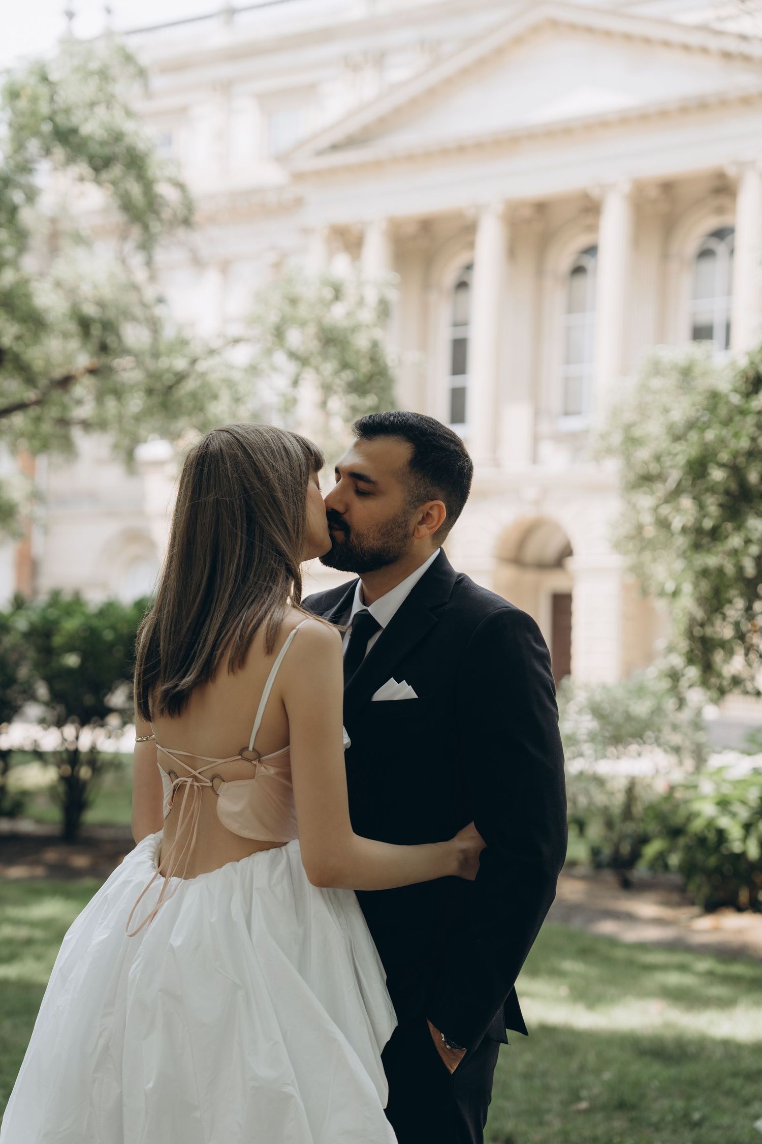 The couple share a kiss in front of a white mansion with classic Greek revival architecture on their wedding day.