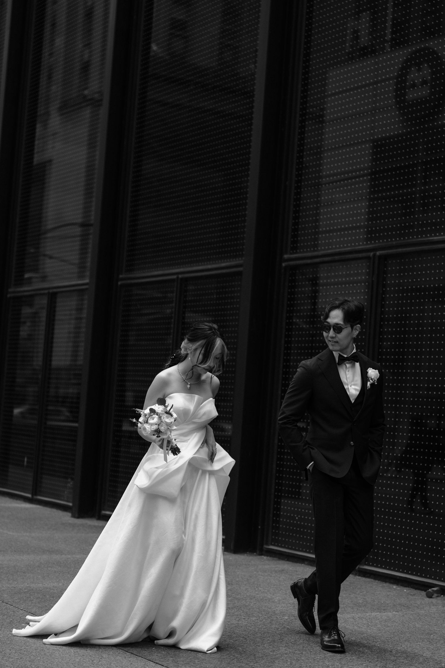 A bride in a flowing white gown and groom in a black tuxedo walk on a city street in black and white photography.