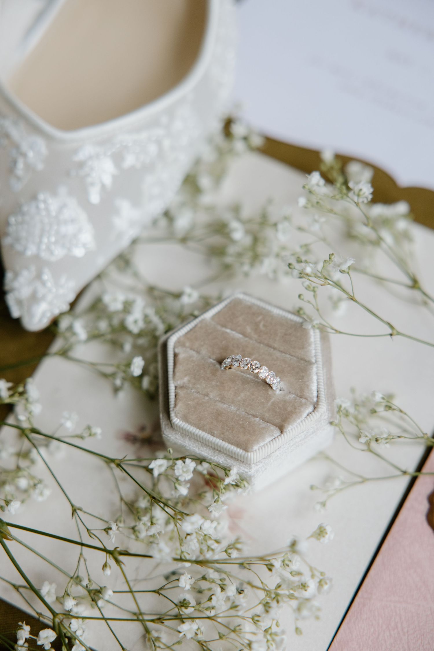 Wedding accessories including a beaded white bridal shoe, delicate baby's breath flowers, and diamond ring in hexagonal box.
