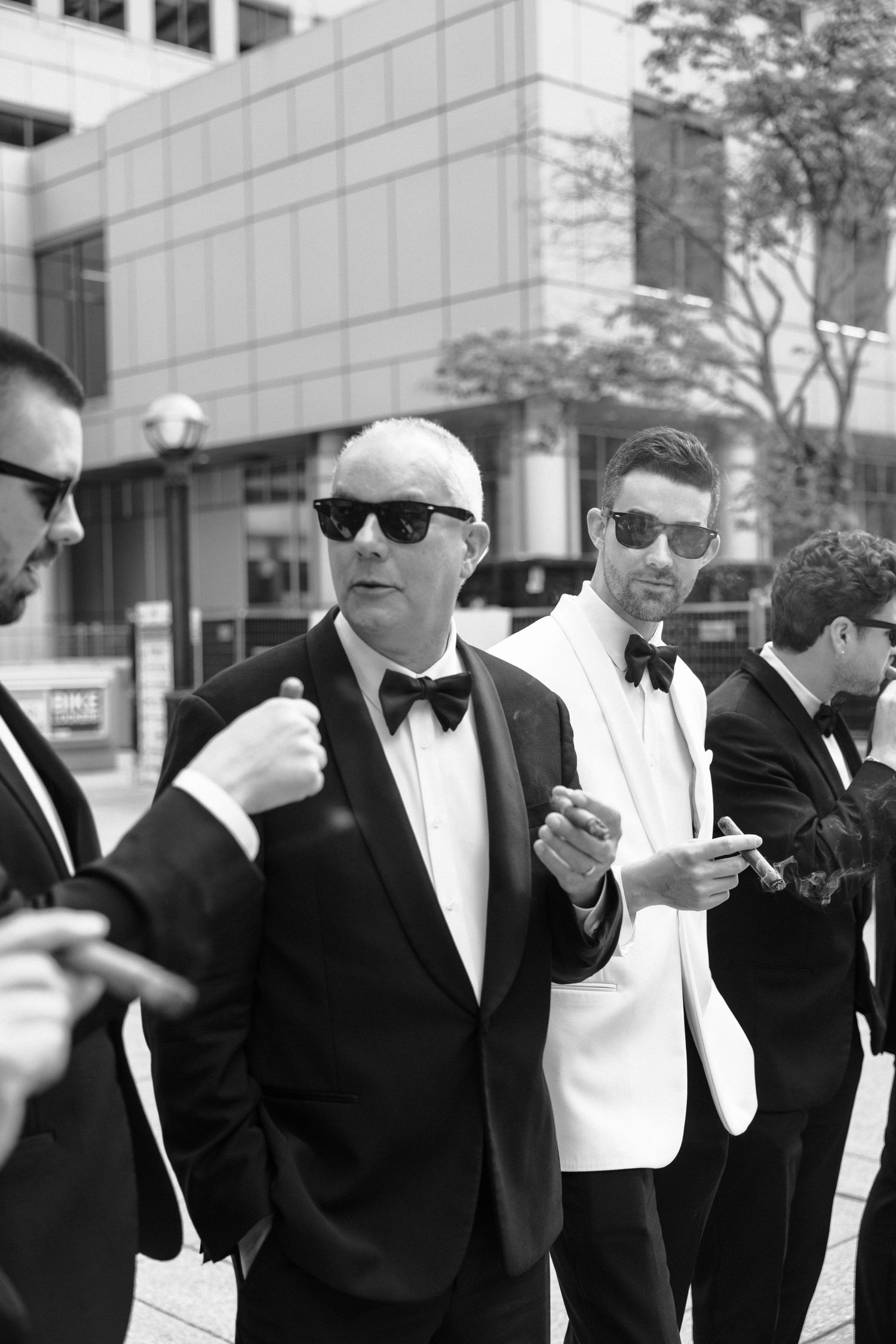 A group of well-dressed men in formal black and white tuxedos and sunglasses stand together on a city street.