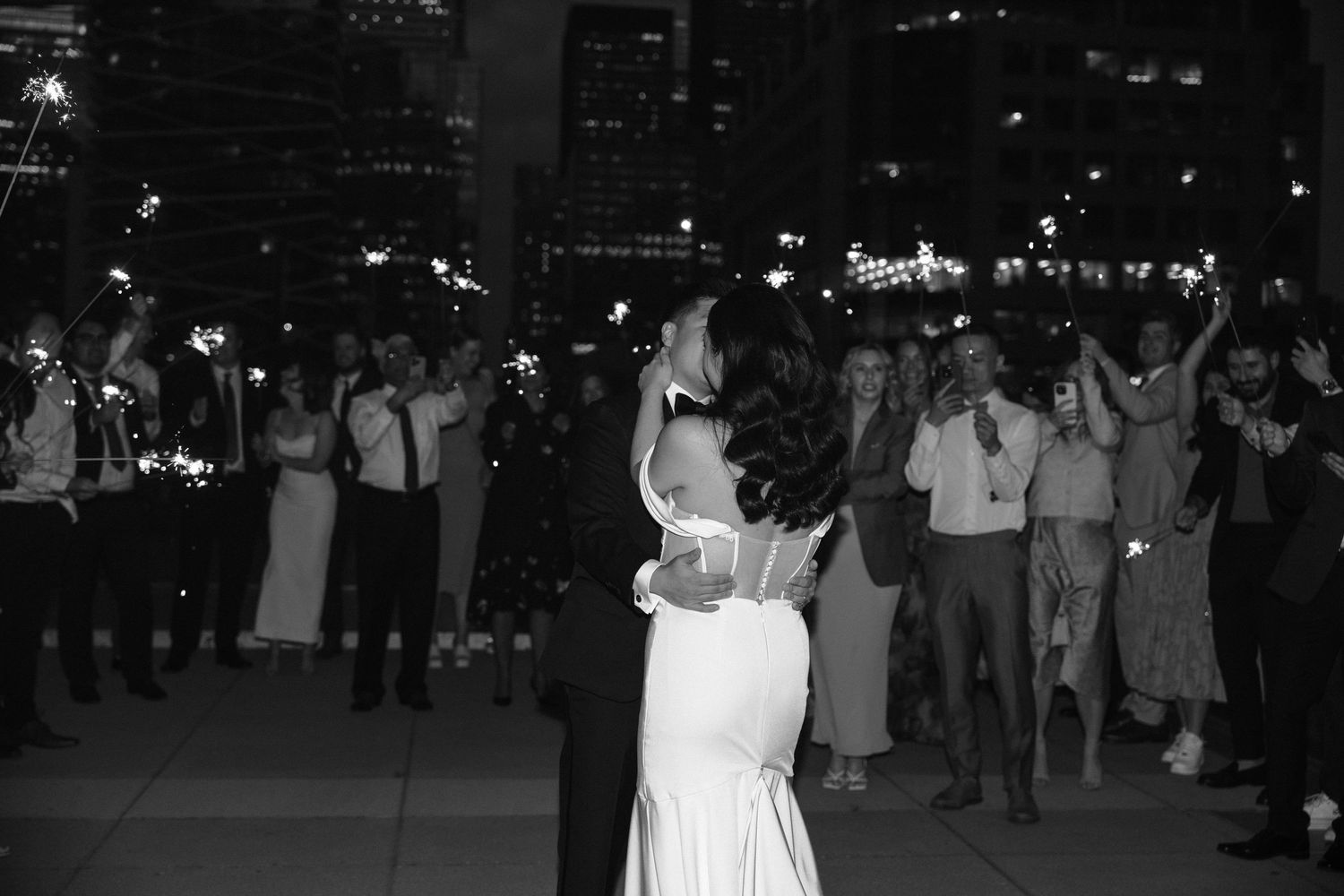 Black and white photo of a couple slow dancing at a nighttime wedding reception with onlookers in the background.