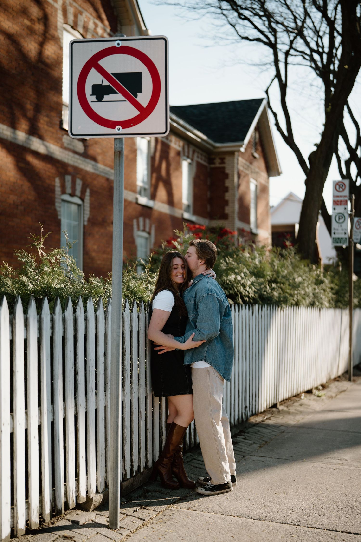 A couple embraces on a sidewalk next to a white picket fence and a no-truck traffic sign on a sunny afternoon.
