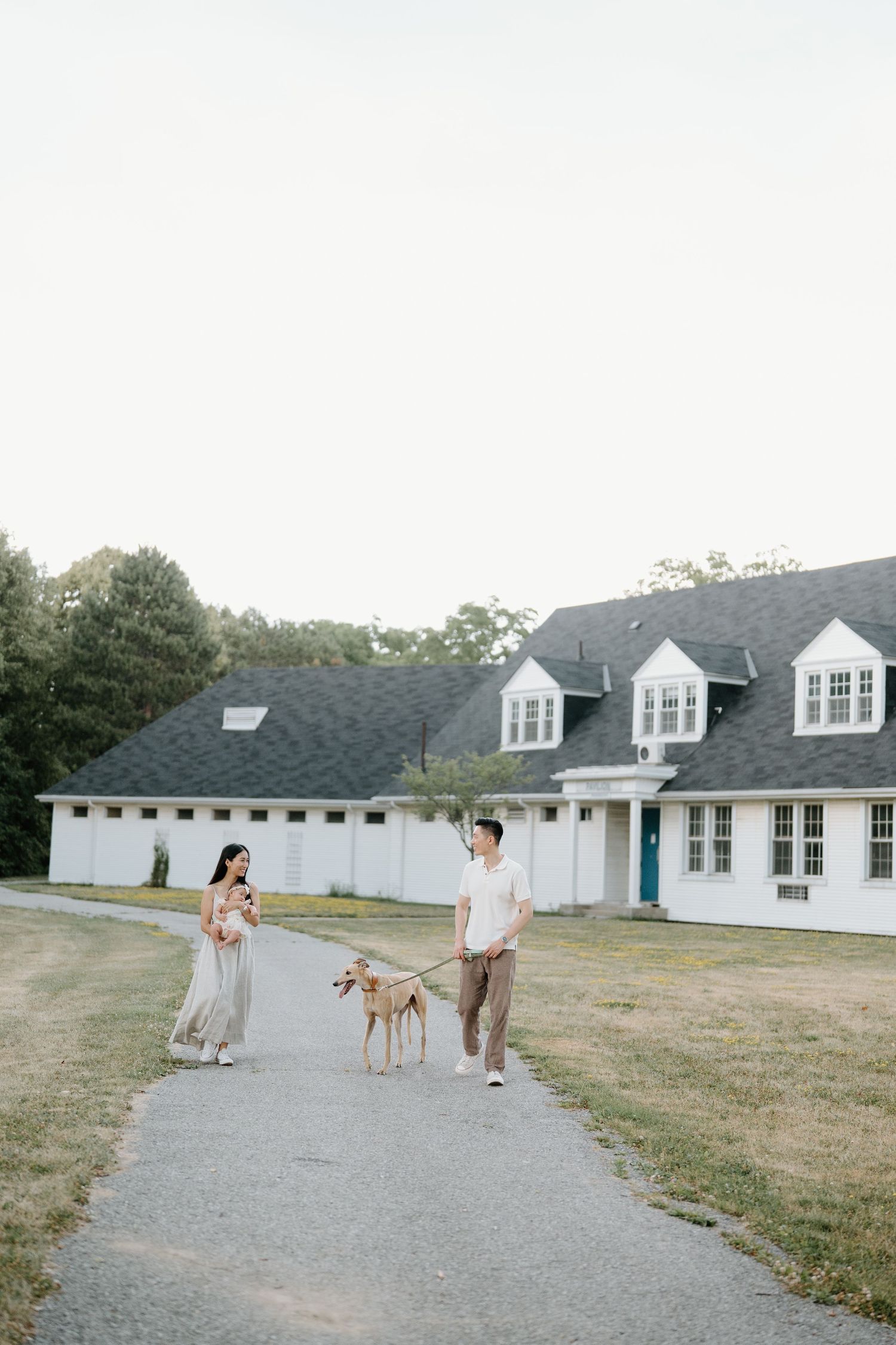 A couple walks their dog on a path in front of a white Cape Cod style house with dormer windows.
