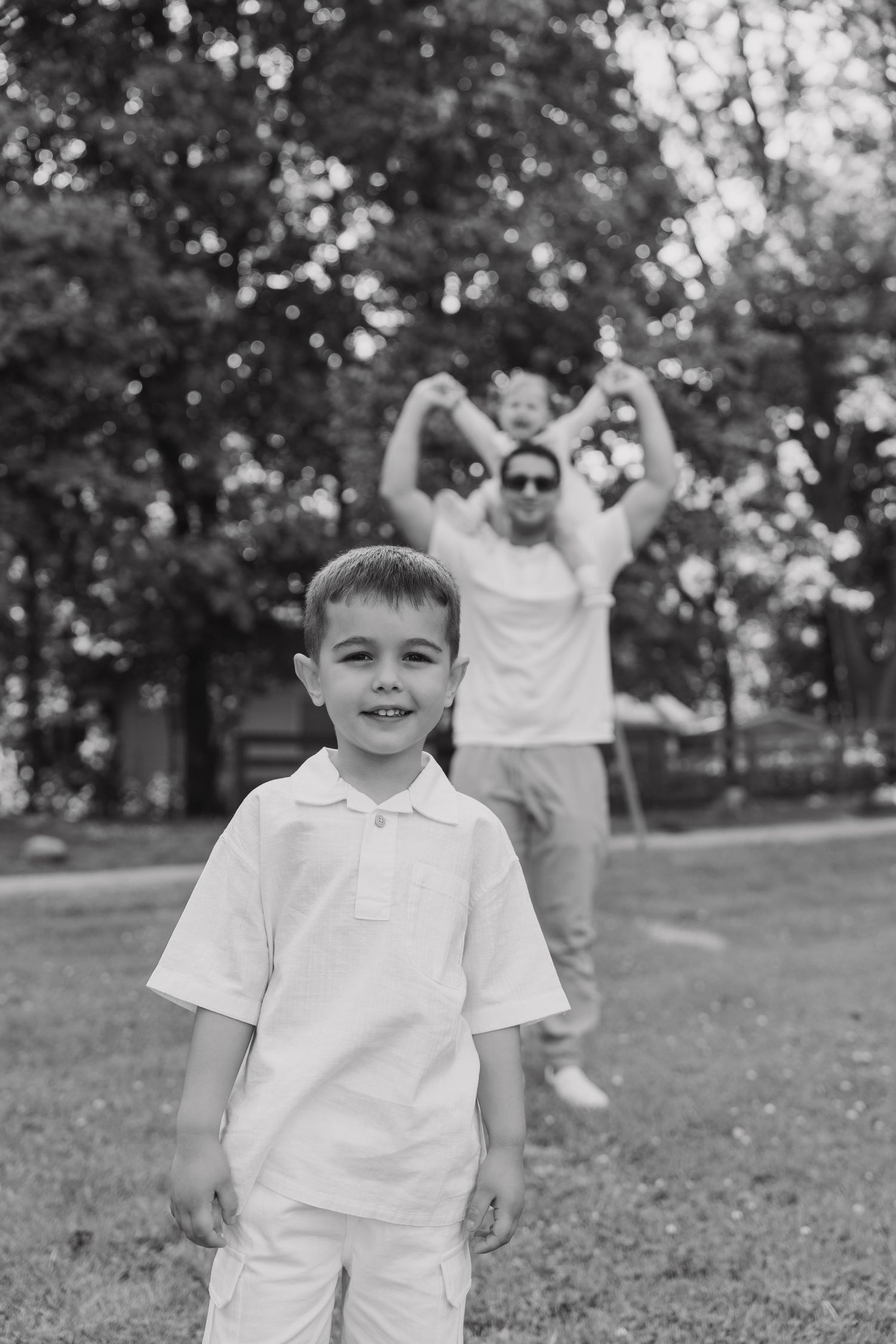 Black and white photo of a young boy in a white outfit standing in a grassy area with someone making a heart gesture behind him.